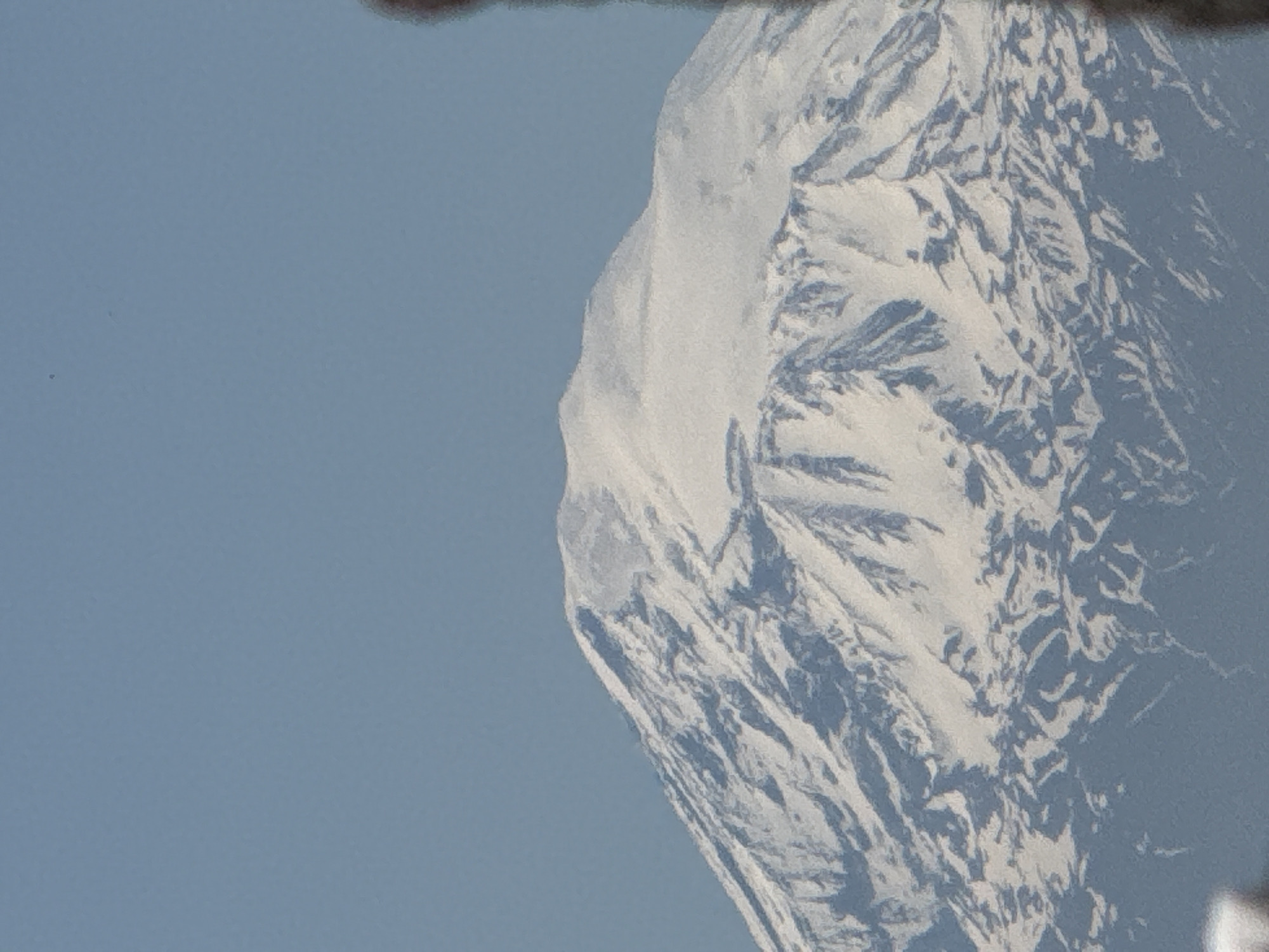 This media file features a majestic, snow-capped mountain peak set against a clear blue sky. The image appears to be rotated clockwise, positioning the mountain on the right side of the frame, extending from the bottom right corner towards the top right. The left side of the image is dominated by a uniform, pale blue sky.
The mountain's surface is rugged and heavily textured, covered in vast expanses of brilliant white snow and ice, interspersed with darker, possibly rocky regions and deep shadows that delineate its complex geological formations. These shadows suggest that the mountain is illuminated by sunlight, highlighting its peaks, ridges, and crevices. The snow cover is thick, indicating either a high altitude, a cold climate, or both.
There are no people or man-made objects visible in the scene. No specific activity or event is taking place; the image captures a still landscape. The time of day appears to be daytime, given the bright, clear blue sky and the distinct shadows on the mountain. There are no discernible weather phenomena such as clouds, suggesting fair conditions. No visible text is present in the image. The overall setting is a grand mountainous environment.