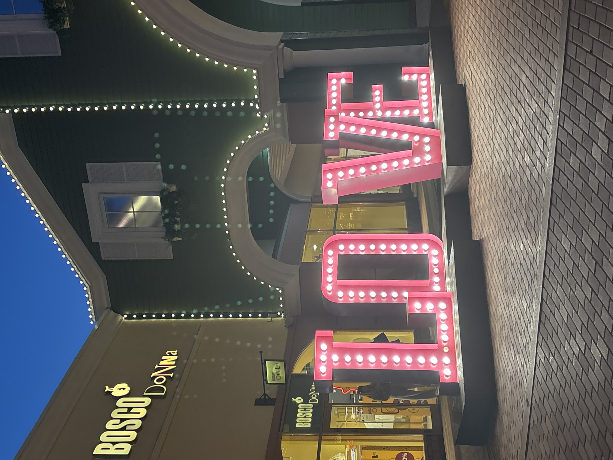 An outdoor scene at dusk shows a large, illuminated pink sign spelling "LOVE" in front of buildings adorned with strings of white lights. The "LOVE" sign is positioned on a paved walkway. To the left, a building with a sign reading "BOSCO Donna" is visible, with storefront windows displaying merchandise. The architecture suggests a commercial or shopping area, possibly a mall or outdoor retail complex. The sky is a deep blue, indicating twilight.