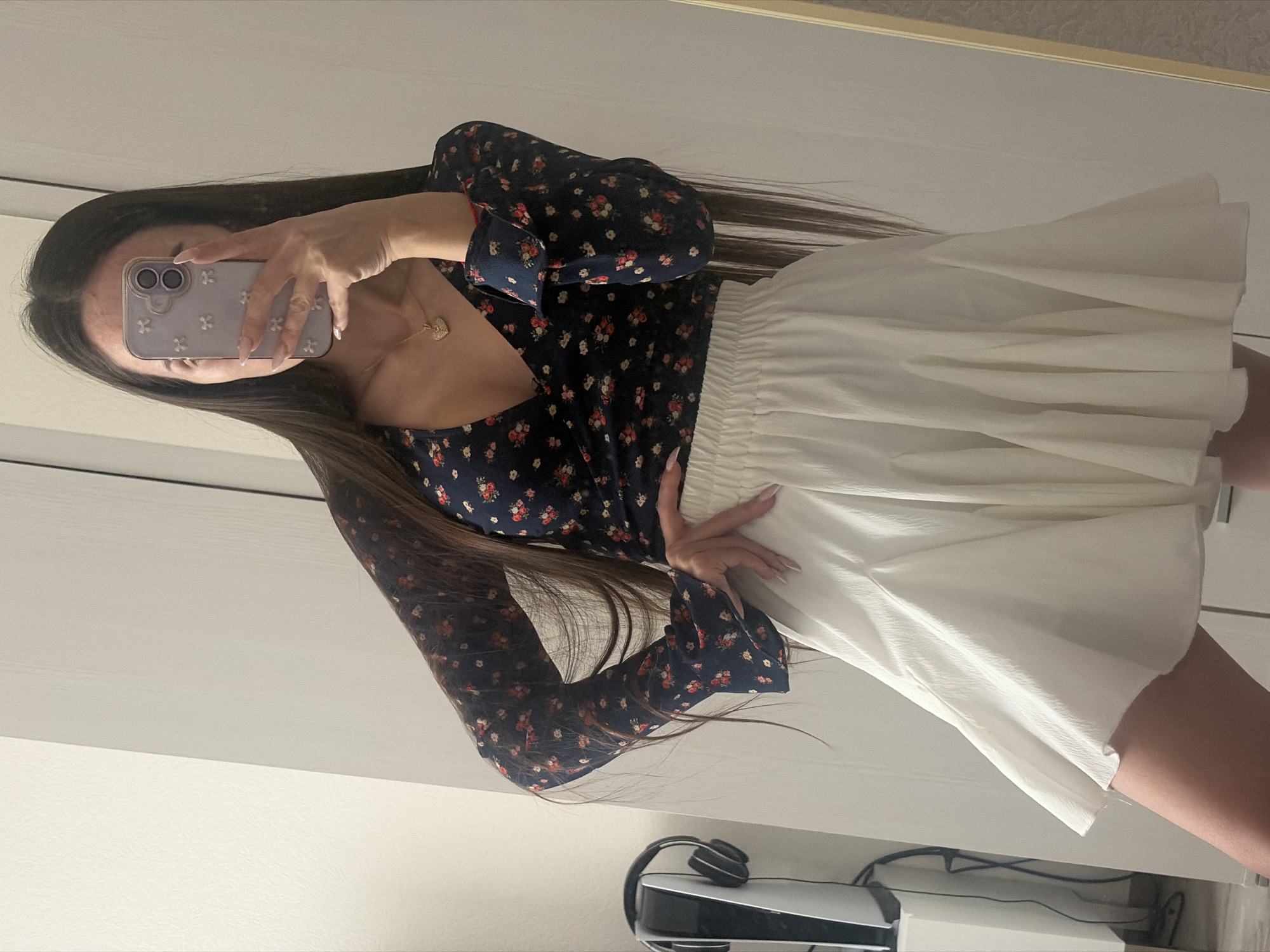 An indoor mirror selfie captures a person, likely female, with long dark hair. Her face is obscured by a light purple smartphone with a white floral-patterned case, which she holds in her right hand. She wears a dark blue long-sleeved top adorned with a red and white floral print, paired with a light cream or off-white pleated mini-skirt. A gold chain necklace with a heart-shaped pendant is visible around her neck. Her nails are long and manicured with a light polish.
The setting appears to be a private room, possibly in Saint Petersburg, Russia, featuring light-colored walls or furniture in the background. In the lower right corner of the reflection, a white and black PlayStation 5 console is visible, with black headphones resting on top of it and various cables behind it. The image is oriented horizontally, showing the person standing sideways to the camera.