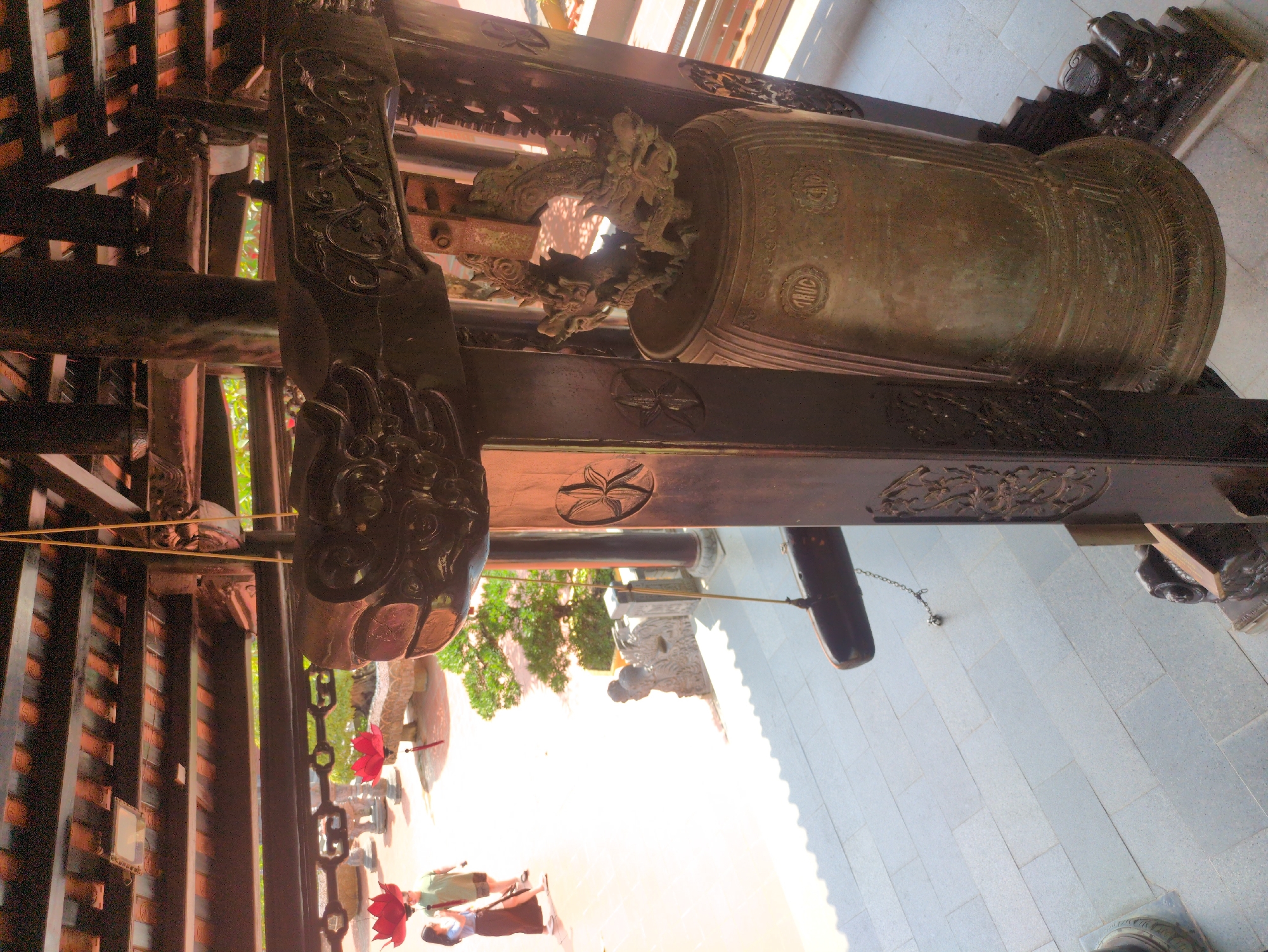 The image shows a large bronze bell mounted within a wooden framework, likely at a Buddhist temple in Phú Quốc, Vietnam. The framework is intricately carved with decorative elements. The bell is positioned horizontally, with its opening facing away from the viewer. A wooden striker hangs nearby. The scene is partially outdoors, with glimpses of people, foliage, and a tiled courtyard.