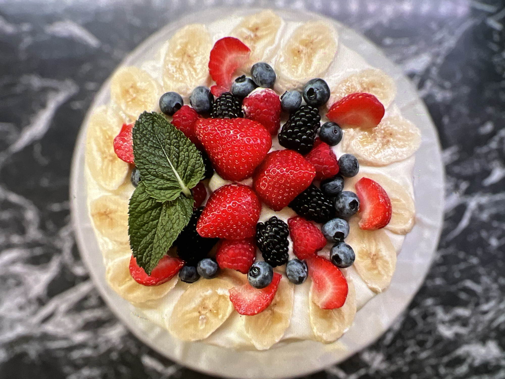 A close-up, overhead shot shows a white cake topped with various fruits and a sprig of mint. Slices of banana are arranged around the perimeter of the cake, and the center is adorned with whole strawberries, raspberries, blueberries, and blackberries. A sprig of fresh mint sits atop the arrangement of berries. The cake rests on a white plate, which is on a dark marble surface. The image is bright and clear, highlighting the vibrant colors of the fruit. The location is not specified within the image.