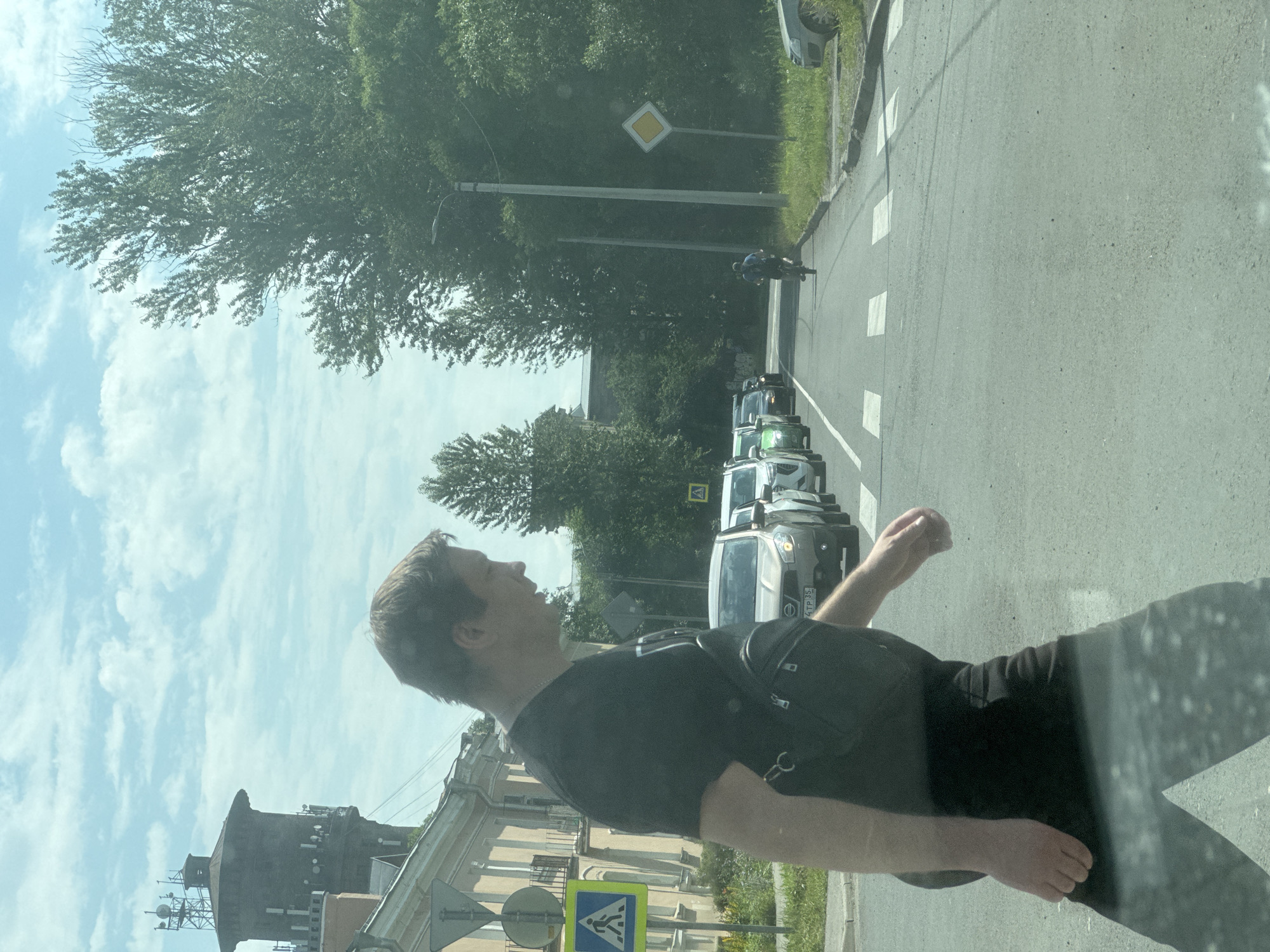 A man in a black t-shirt and black pants walks across a crosswalk in Metallostroy, Russia. He is wearing a black crossbody bag. In the background, there are several cars stopped at the crosswalk, and a cyclist is riding down the street. There is a water tower with antennas on top in the distance. The sky is blue with some clouds.