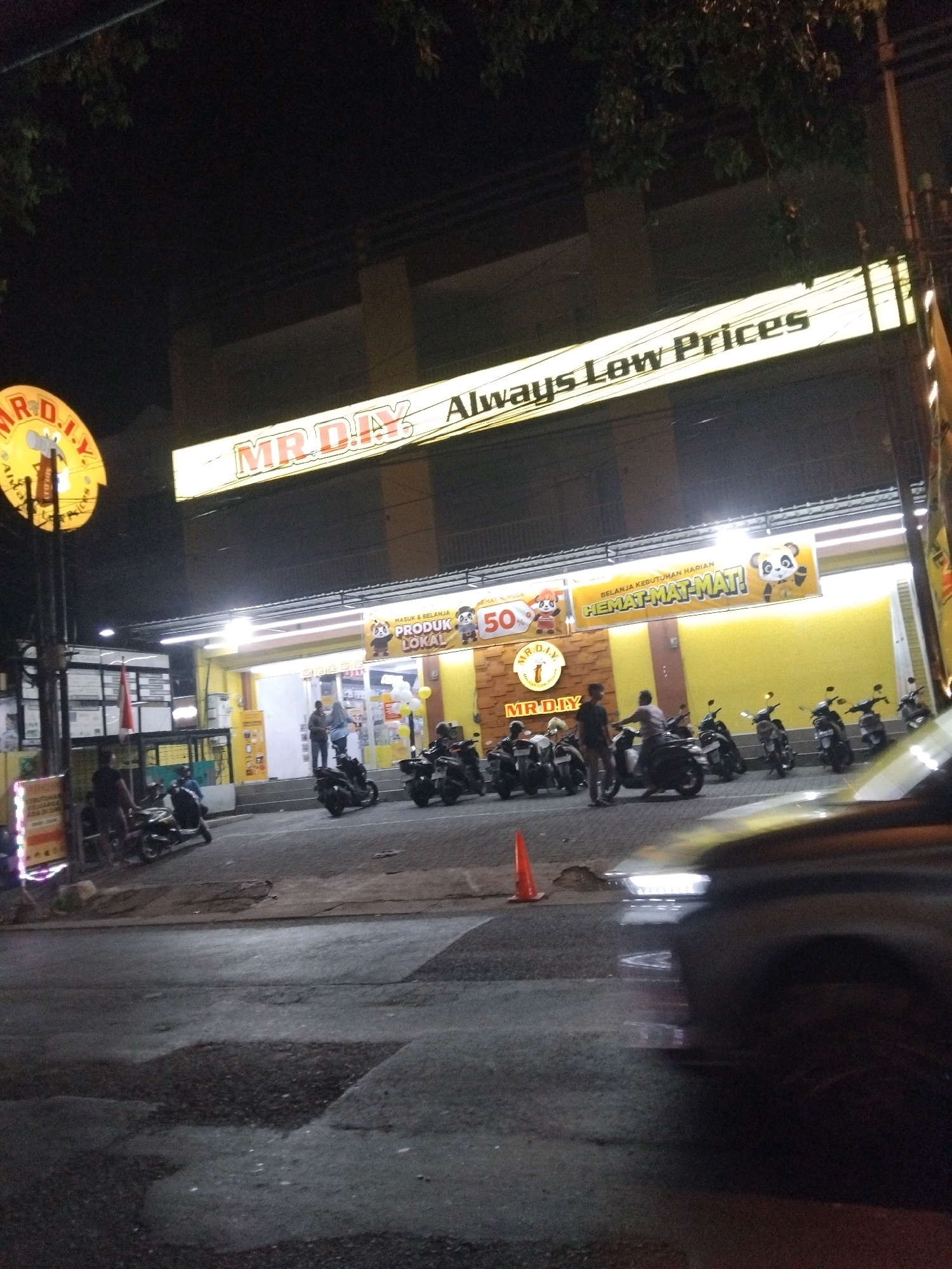 The image shows a night scene in front of a MR. DIY store located in Wedoro, Indonesia. The store's prominent yellow signage reads "MR. DIY Always Low Prices". The facade of the store is brightly lit, and a banner with cartoon pandas and the text "HEMAT-MAT-MAT!" is displayed above the entrance. Several motorcycles are parked outside the store, and a few individuals are standing or walking near them. A bright orange traffic cone is visible on the pavement. A portion of a dark-colored car, possibly in motion, is reflected in the foreground. The street is paved with asphalt, and some areas show signs of wear and tear. Trees with dark foliage are visible against the night sky.