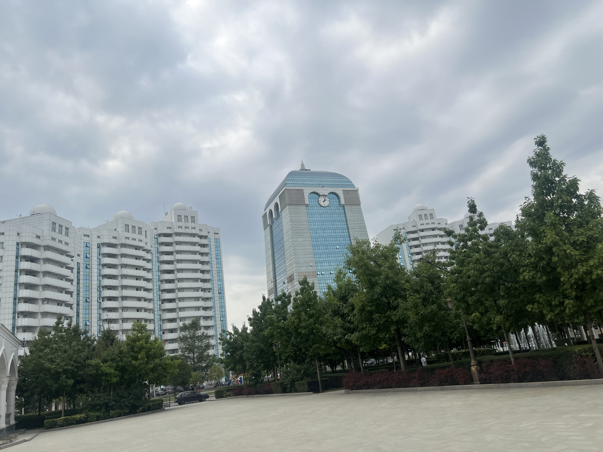 The image depicts the city center of Shali, Russia, under an overcast sky. In the foreground, a wide, light-colored paved area, possibly a plaza or walkway, dominates the lower half of the frame. To the left, a building with white facade and numerous balconies can be seen. This building is a residential complex. In the middle ground, a tall, modern skyscraper with a glass facade and a distinctive arched top featuring a clock is prominent. Two similar residential complexes are visible flanking the central skyscraper. A row of lush green trees with dark red bushes lining the bottom of the planter bed runs across the middle ground, separating the paved area from the buildings and trees. In the background, several parked cars are visible. The sky is filled with gray clouds.