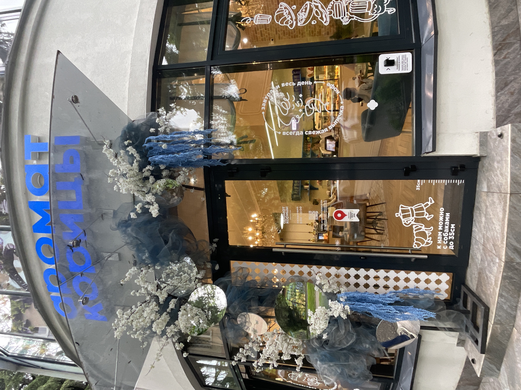 The image shows the entrance to "Аромат Корицы" (Aroma of Cinnamon), a cafe located in Sochi, Russia. The storefront is decorated with white blossoms and blue floral accents. Glass doors showcase the interior of the cafe. Lettering on the glass doors advertises "fresh pastries".
---