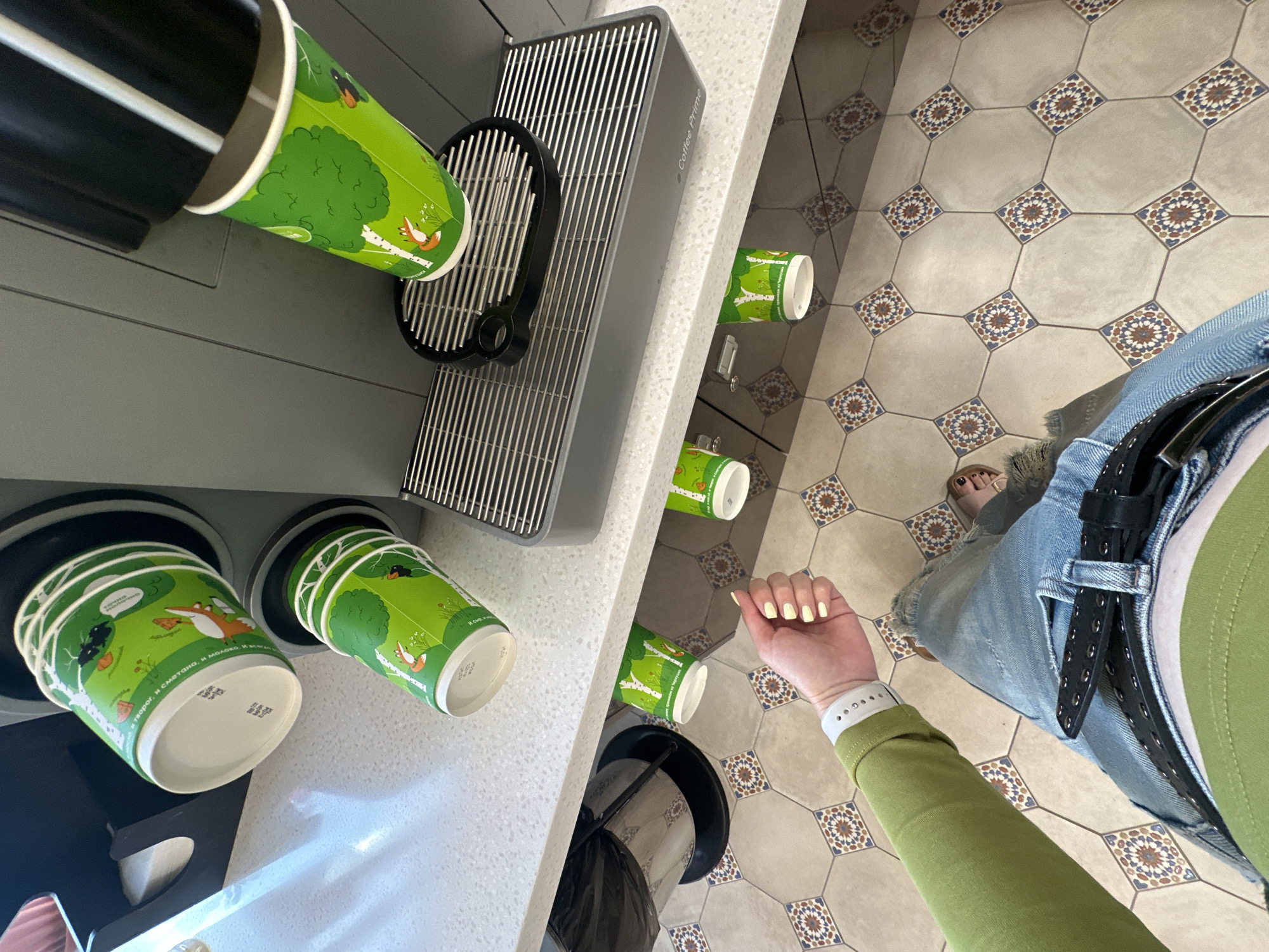This is a first-person perspective shot taken from a standing position, looking down at a coffee machine and surrounding counter area. The scene appears to be in Perm, Russia, based on text on the coffee cups.
In the foreground, a person's left arm, clad in a lime-green long-sleeved shirt, is visible, along with their hand. The person is wearing a white band on their wrist, possibly a watch or fitness tracker. They are also wearing light blue denim jeans, a black belt with silver grommets, and a green top. Their feet are visible at the bottom of the frame, wearing open-toed sandals, with painted toenails.
The main subject of the image is a coffee machine with multiple green paper cups featuring a forest-themed design with foxes and birds. One cup is positioned under the machine's dispenser, actively receiving a beverage. Other cups are visible in dispensers on the counter and below the counter. The counter is white and speckled, and the floor is tiled with an octagonal pattern in beige, blue, and red. To the left of the coffee machine, a black trash receptacle is partially visible. The image is taken from a high angle, looking downwards towards the coffee machine and the floor.