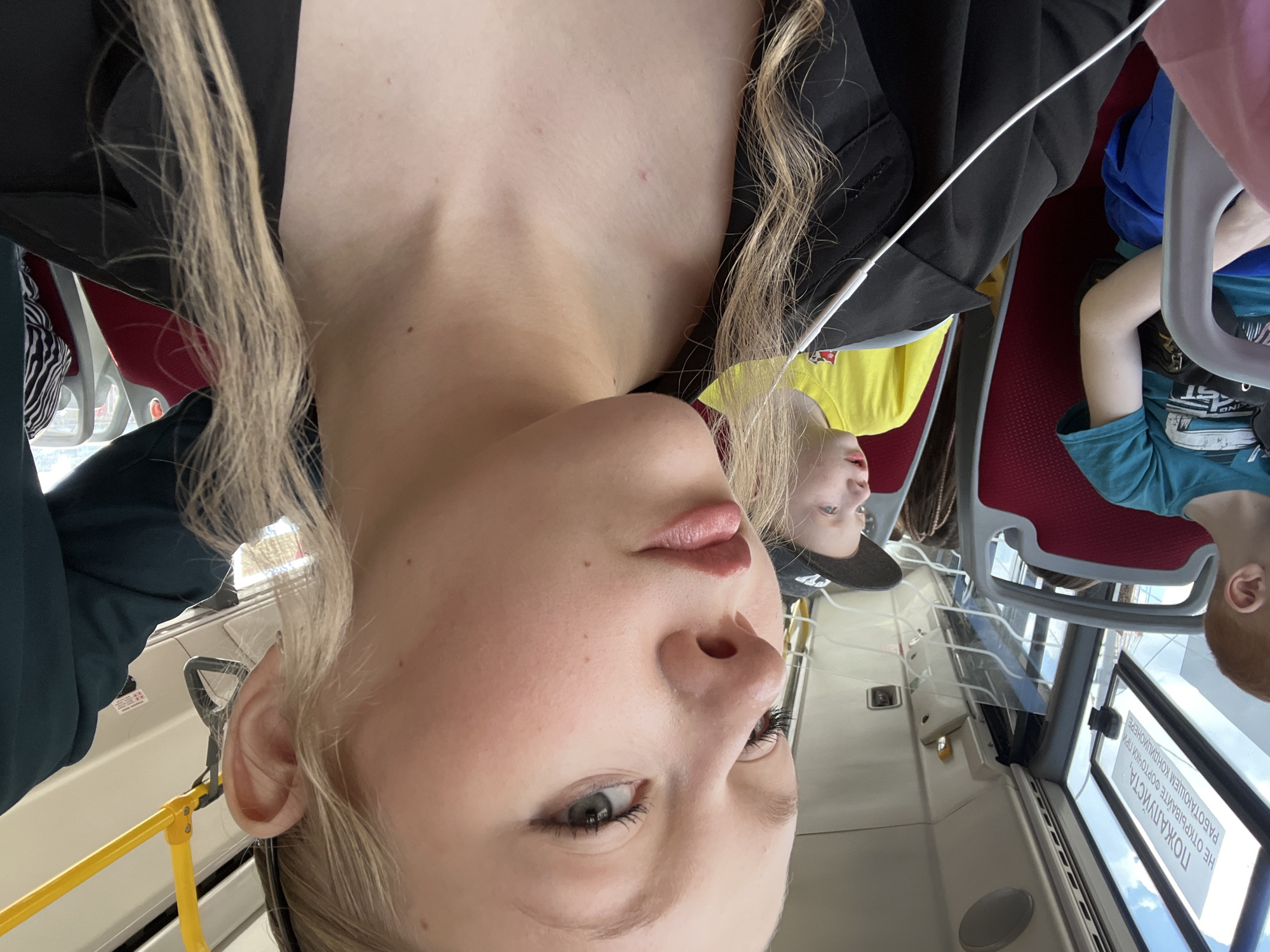 The image shows a young woman with blonde hair, wearing a black jacket and earbuds, taking a selfie on a bus. She is looking towards the camera with a neutral expression. In the background, two young boys are visible, one in a blue shirt and the other in a yellow shirt. The bus interior is depicted with red seats and a yellow handrail. A sign in Russian is visible on the window, partially obscured. The scene appears to be in the city of Perm, Russia, based on the presence of Russian text on the sign.