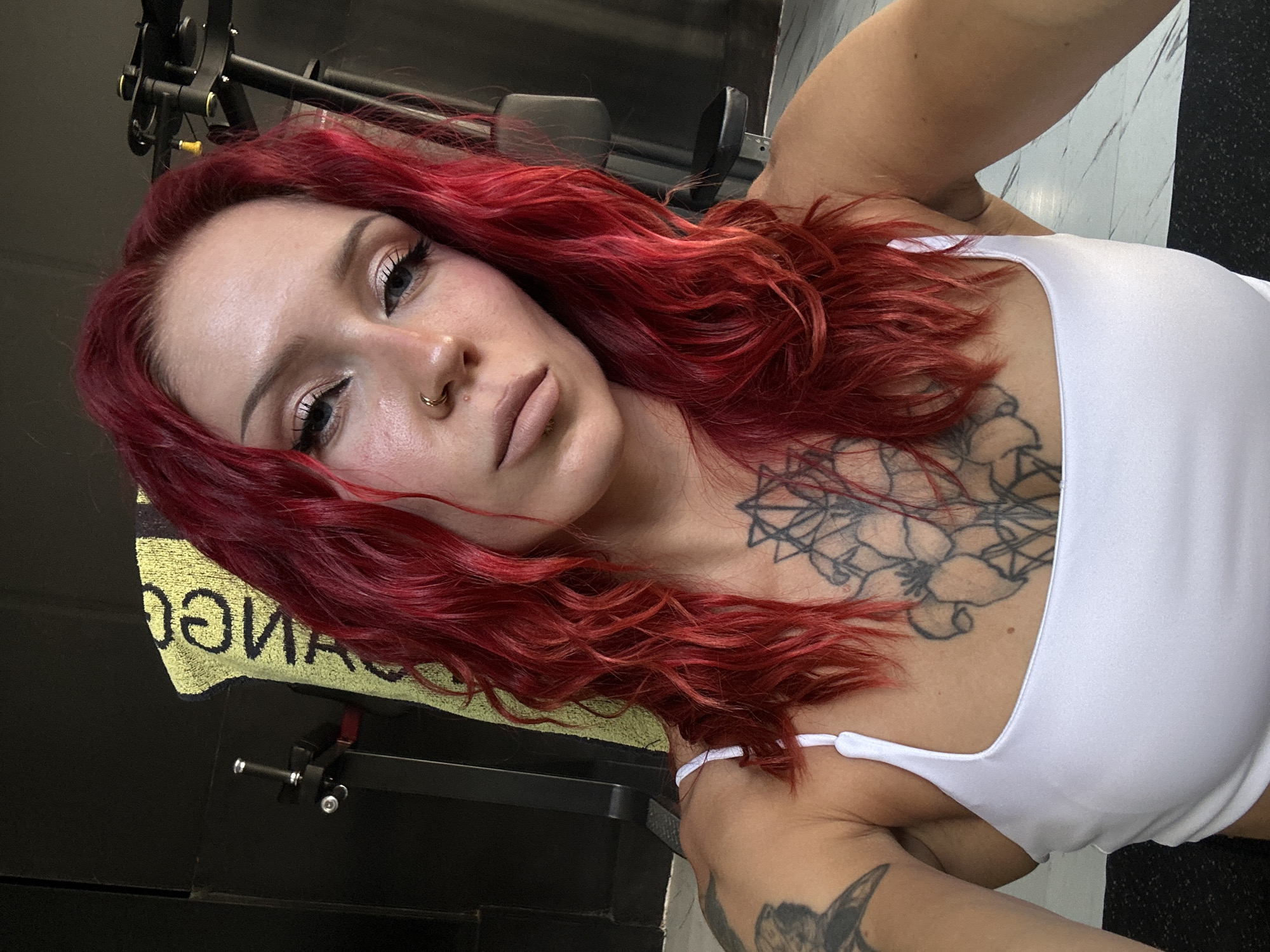 A medium shot, taken from a slightly low angle, shows a woman with bright red, wavy hair. She is wearing a white tank top and has a tattoo on her chest. The woman has blue eyes, winged eyeliner, and a nose ring. She is looking directly at the camera. In the background, there is a yellow towel with text on it, a piece of gym equipment, and a wall. The floor has a marble pattern. The setting appears to be a gym.