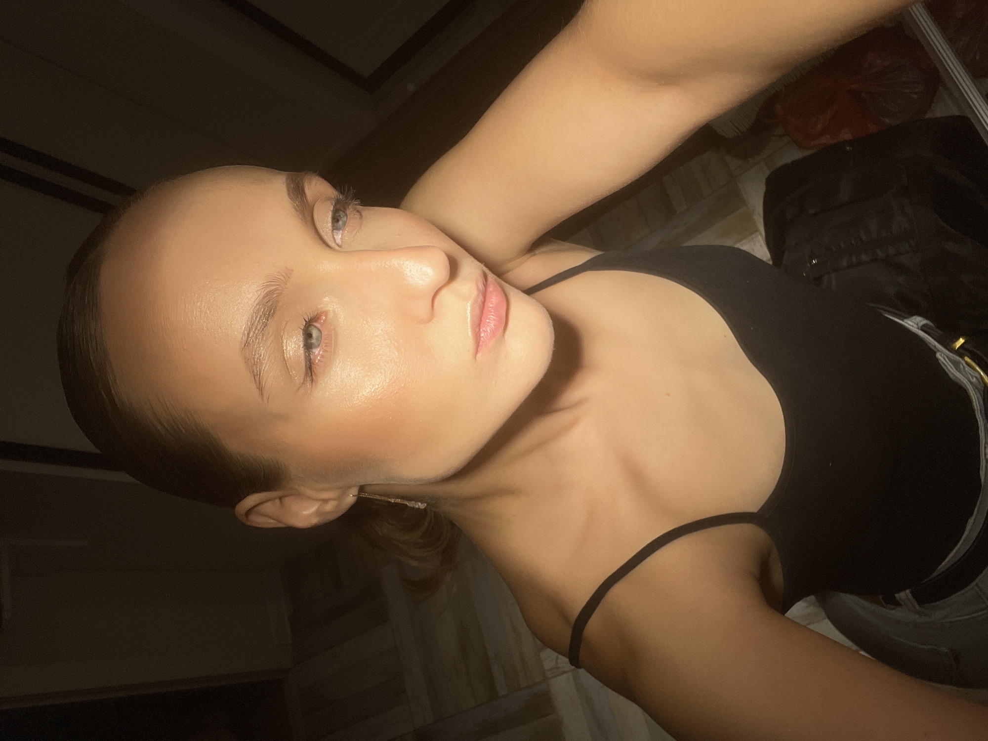 An overhead, close-up shot captures a fair-skinned woman from the chest up. Her hair is pulled back tightly, revealing her forehead and ears. She wears a black tank top and a thin gold earring. Her eyes are open and looking to the right, with light makeup on her eyelids. The lighting suggests an indoor setting with a single light source creating strong shadows and highlighting the woman's cheekbones, nose, and forehead. The background is dark, with faint patterns of what appears to be a floor or wall visible. There are no discernible Chaikovsky, Russia landmarks.