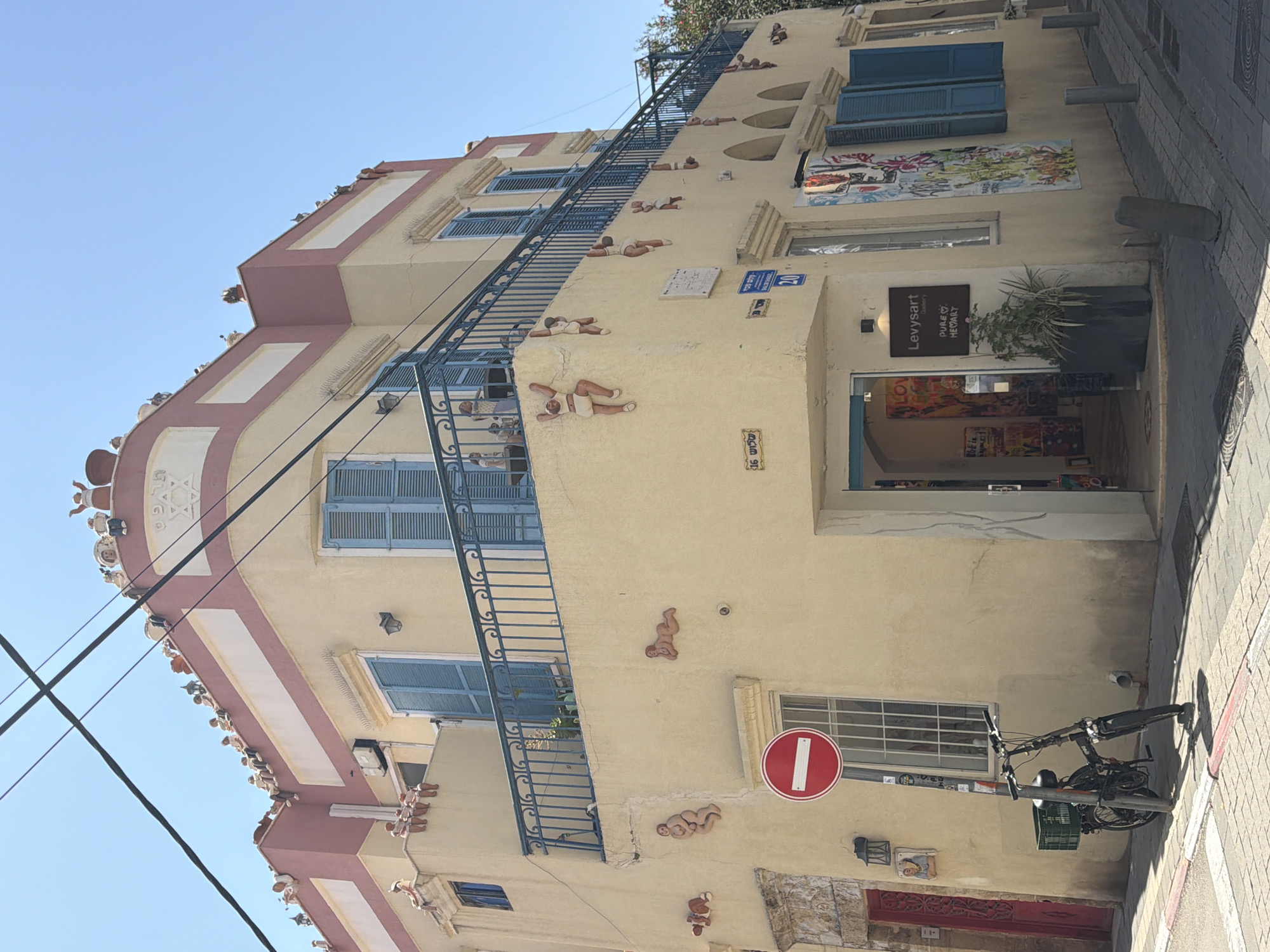 The image captures a view of a unique building in Tel Aviv, Israel, known for its distinctive architectural features and artistic embellishments. The structure is characterized by a pale yellow facade, accented with pink trim and adorned with numerous small sculptures of reclining human figures scattered across its surface. A blue metal external staircase ascends the building's front, adding to its visual interest.
The building's facade also features blue-shuttered windows, and artwork displayed prominently beside an open doorway that leads into a space identified by a sign as "Levysart." A bicycle is parked in front of the building. A traffic sign is mounted on the wall next to the window. The sky is clear. The building is located on a paved street.