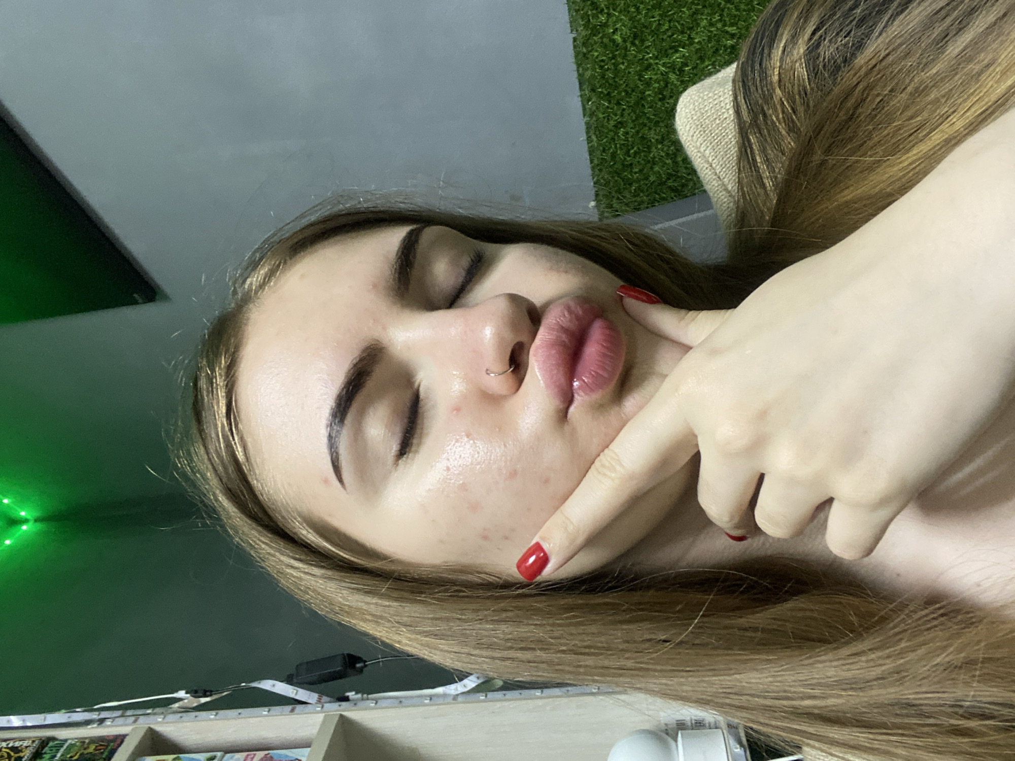 A close-up portrait shows a young woman with her eyes closed and lips puckered in a pouting expression, with her right index finger gently pressed against her cheek. Her fingernails are painted a vibrant red. She has long, light brown hair with blonde highlights and a small silver hoop nose ring in her left nostril. Her skin is fair, with some visible blemishes on her face.
The scene is indoors, with various decorative and functional elements in the background. To the upper right, a section of green artificial turf is visible next to a beige, woven fabric or cushion. On the left side, a strong green light emanates, possibly from an LED strip, reflecting on the adjacent dark surface. Towards the bottom, a white LED strip light with a black connector is affixed to a light-colored wooden shelf or frame. Below this, multiple shelves hold items that resemble books, although any text on them is illegible due to the distance and angle. The lighting appears to be artificial, suggesting an indoor setting, possibly at night or in a dimly lit room. The woman's pose indicates a playful or stylized moment, potentially a self-portrait.