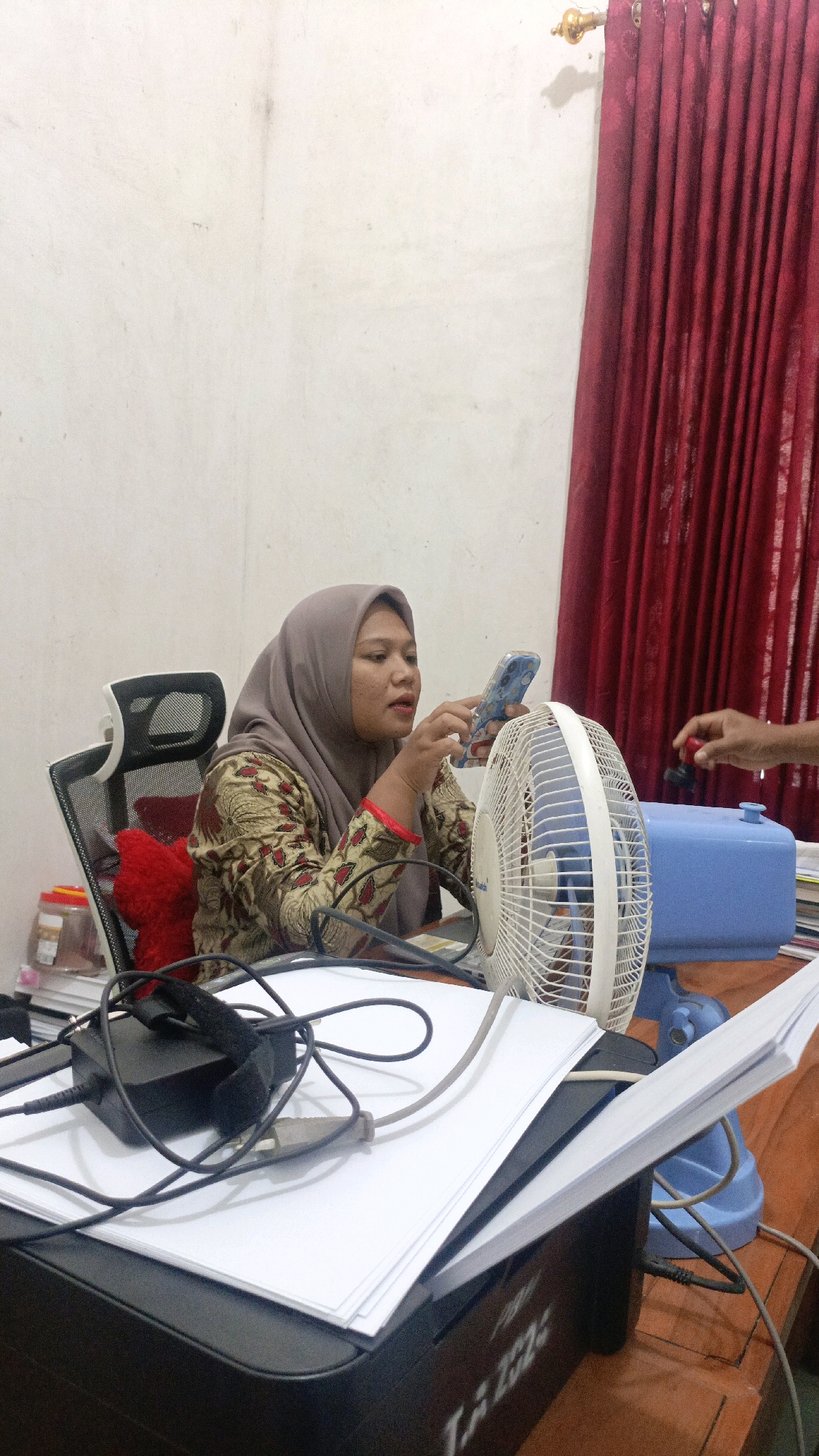 A woman wearing a hijab and patterned clothing sits at a desk, holding a smartphone. A white electric fan is positioned in front of her, and to her right, a hand is shown holding a red stamp above a blue dispenser. The desk is covered with papers, cables, and office equipment. The background features a plain wall and a red curtain. This scene is likely in an office or study space in Taliwang, Indonesia, suggested by the presence of a printer with text that could be Indonesian.