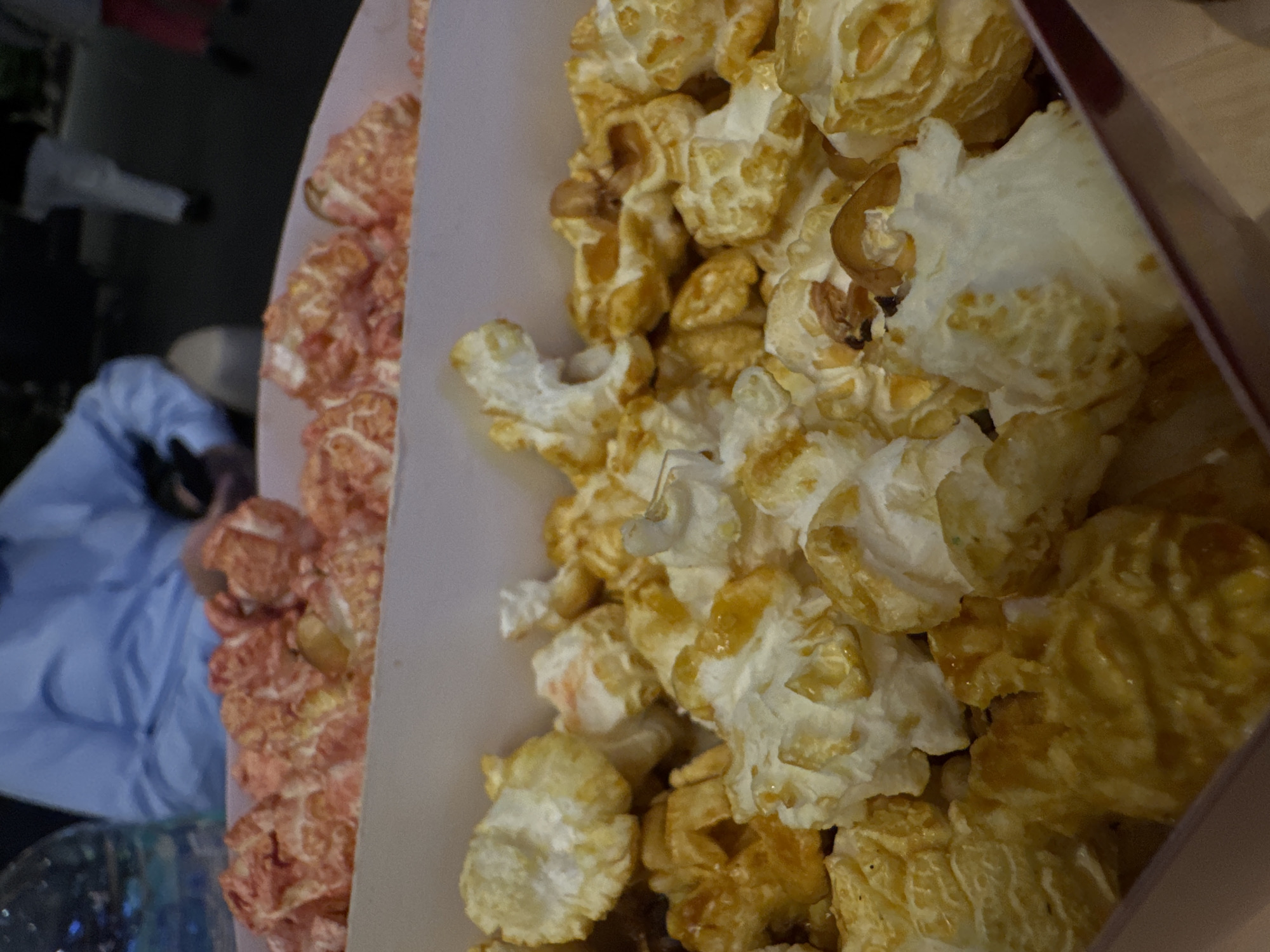 A close-up, tilted, indoor shot of a box of popcorn shows two varieties: one a reddish-pink color on the left and the other a mix of white and golden-brown on the right. Some popcorn pieces have noticeable caramel coating. The box is either made of white cardboard, or lined with white paper.
Blurred in the background on the left is a person in a light blue garment, seated near a glass table or container. The location appears to be indoors, based on the soft lighting and background scene. There is no specific information to connect the image with Mexico City.