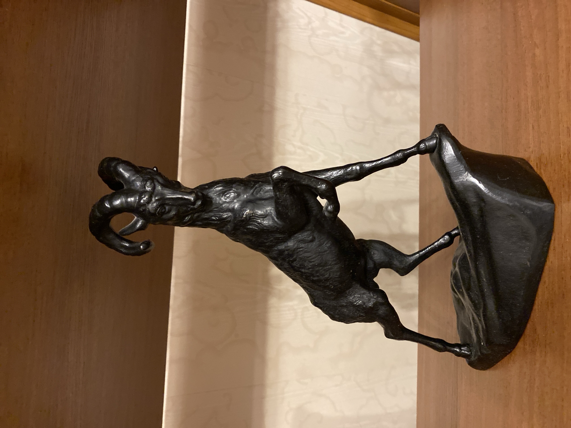 The image displays a dark-colored statue of a bighorn sheep or ram, depicted rearing on its hind legs. The statue, likely made of bronze or cast iron, exhibits prominent, tightly curled horns, a detailed face, and a textured body suggesting fur. Its front hooves are lifted, while its hind hooves are planted on an irregularly shaped, dark-colored base sculpted to resemble rocky terrain. The statue is situated on a polished, flat, medium-brown wooden surface, which appears to be a tabletop or lower shelf. The background consists of a light-beige wall featuring a faint, repeating damask-like pattern. Above this wall, a horizontal segment of a darker brown wooden surface, consistent with an upper shelf, spans the width of the image. A partial view of a lighter wooden structural element is visible on the far right. The scene is set indoors, in an unknown city and country.