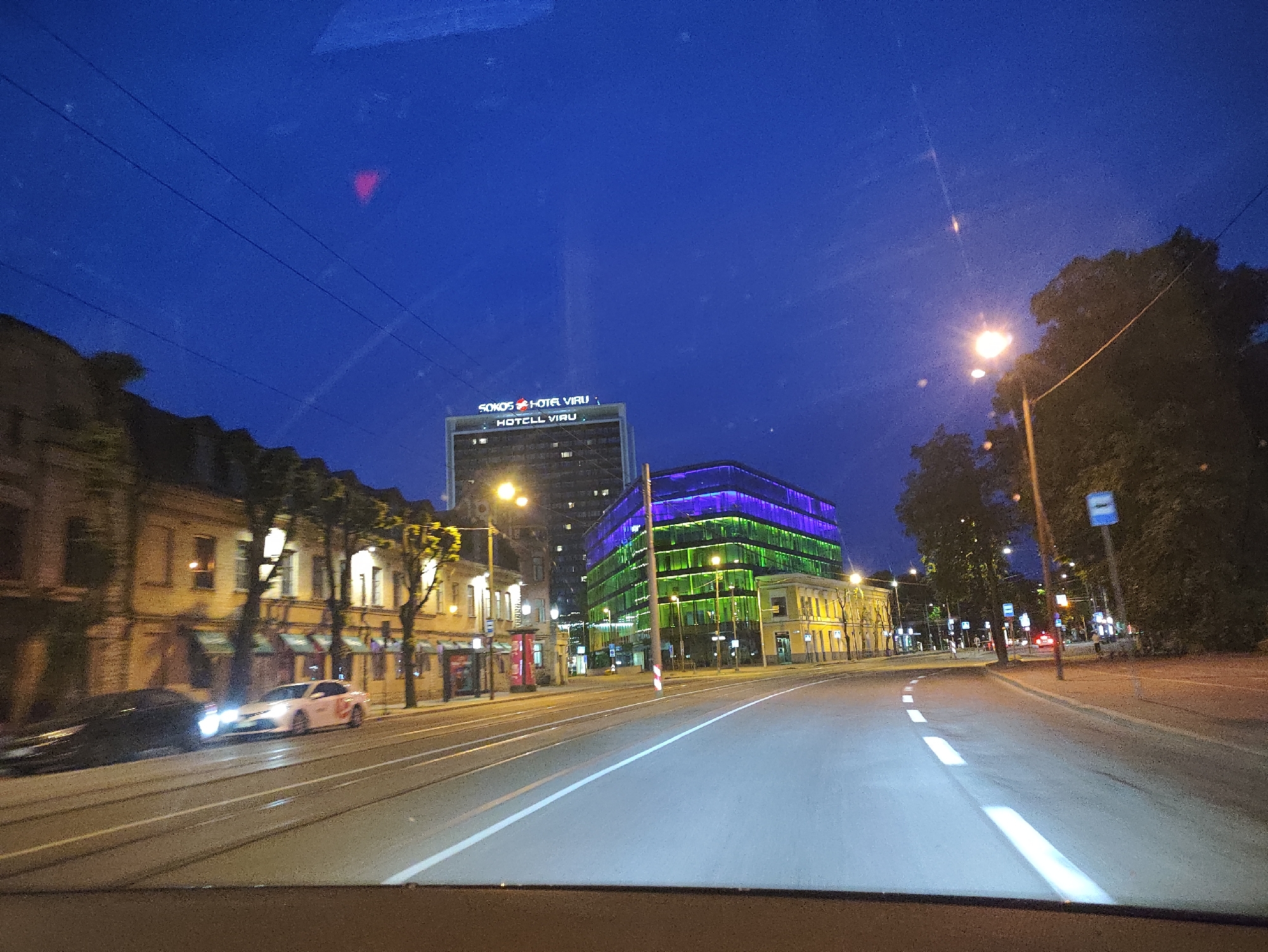 The image shows a street scene in Tallinn, Estonia, at dusk or night. In the foreground, a road with tram tracks and lane markings is visible, with headlights from a car illuminating the right lane. To the left of the road, there are buildings with illuminated windows and trees. Further down the street, a tall, modern building with green and purple lighting is prominent. A sign on top of a smaller, older building reads "SOKOS HOTEL VIRU". Streetlights illuminate the sidewalk and road. The sky is a deep blue, indicating twilight or nighttime.