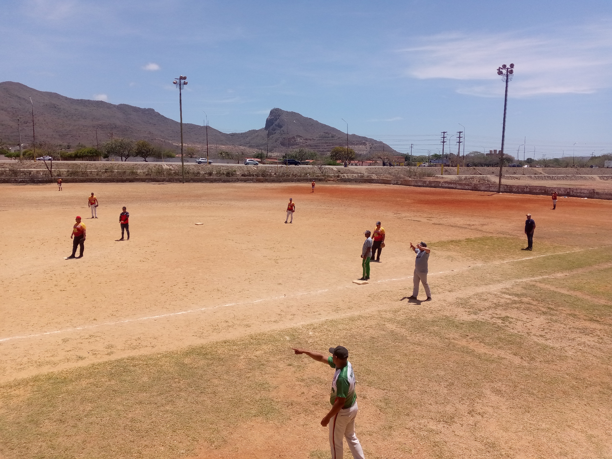 The image depicts a baseball field in what appears to be Macho Muerto, Venezuela, under a clear sky. The field is predominantly dirt, with patches of grass, and is bordered by a low retaining wall. Several individuals are present on the field, some wearing baseball uniforms in varying colors, and others in casual attire. Some individuals are standing still, while others are actively engaged in the game. In the background, there are mountains, trees, roads with vehicular traffic, and light posts. Utility poles and wires are also visible.