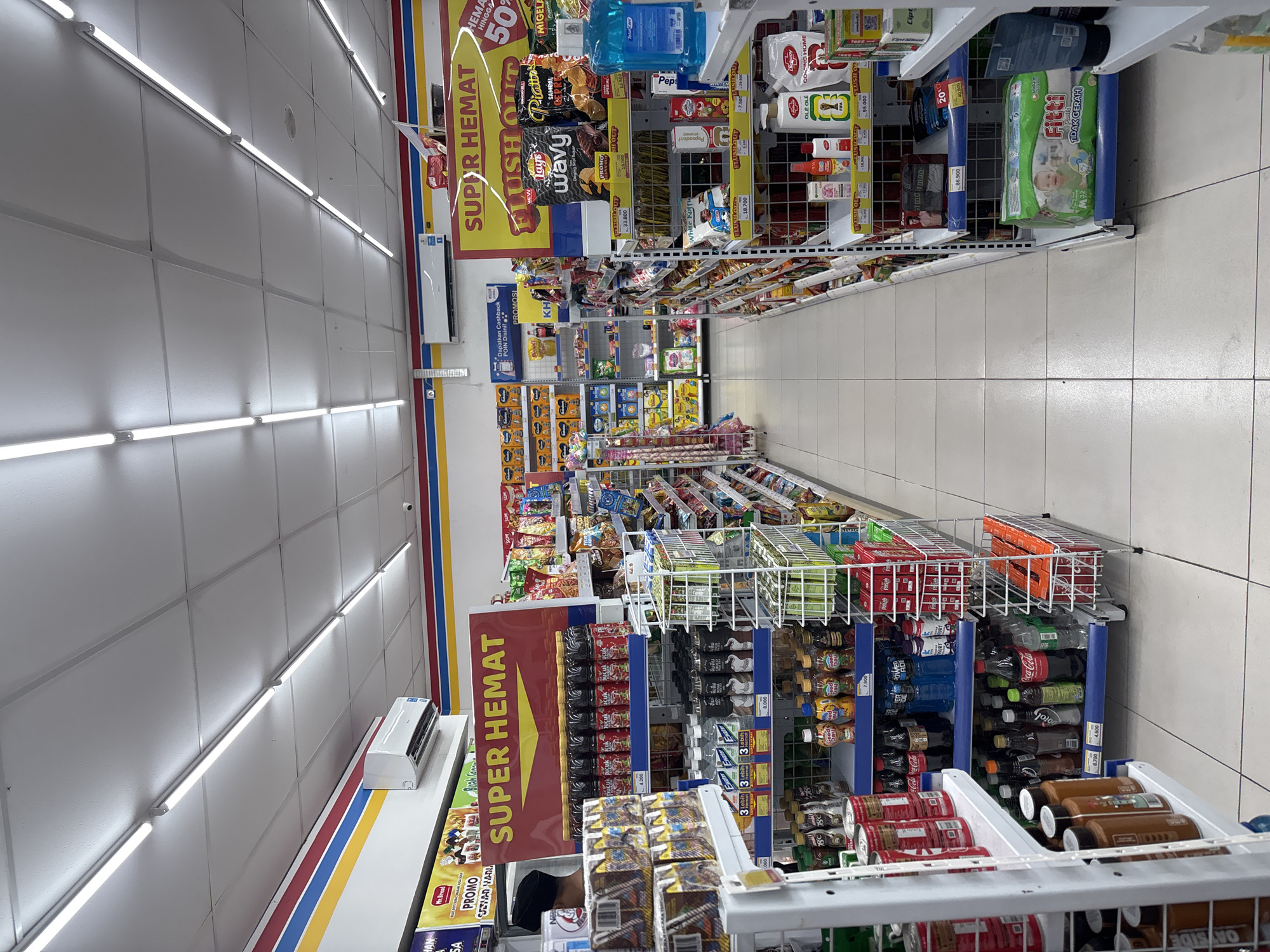 The image depicts the interior of a retail store, likely a convenience store or minimarket, characterized by brightly lit aisles and well-stocked shelves. The store is identified as an Indomaret located in Cirebon, Indonesia, as indicated by signage and product packaging. The perspective is from within an aisle, looking down the length of the store.
In the foreground, on the left, is a display of bottled beverages, including what appears to be tea and soda, with prices displayed on the shelves. Further down this aisle, more shelves are visible, containing various packaged goods such as snacks, drinks, and personal care items. The shelves are organized and labeled with prices and promotional information, like "SUPER HEMAT" (Super Savings).
On the right side of the aisle, a prominent display features branded snacks and drinks, also with promotional signage. This section also includes personal hygiene products and other household items. The flooring appears to be tiled, and the ceiling is fitted with long, fluorescent lighting fixtures.
A few individuals are partially visible. On the left, a person wearing a dark head covering and a light-colored shirt is seen from the back, suggesting they may be an employee or a shopper. The overall environment suggests a typical retail setting focused on convenience and affordability, with an emphasis on promotions and deals.