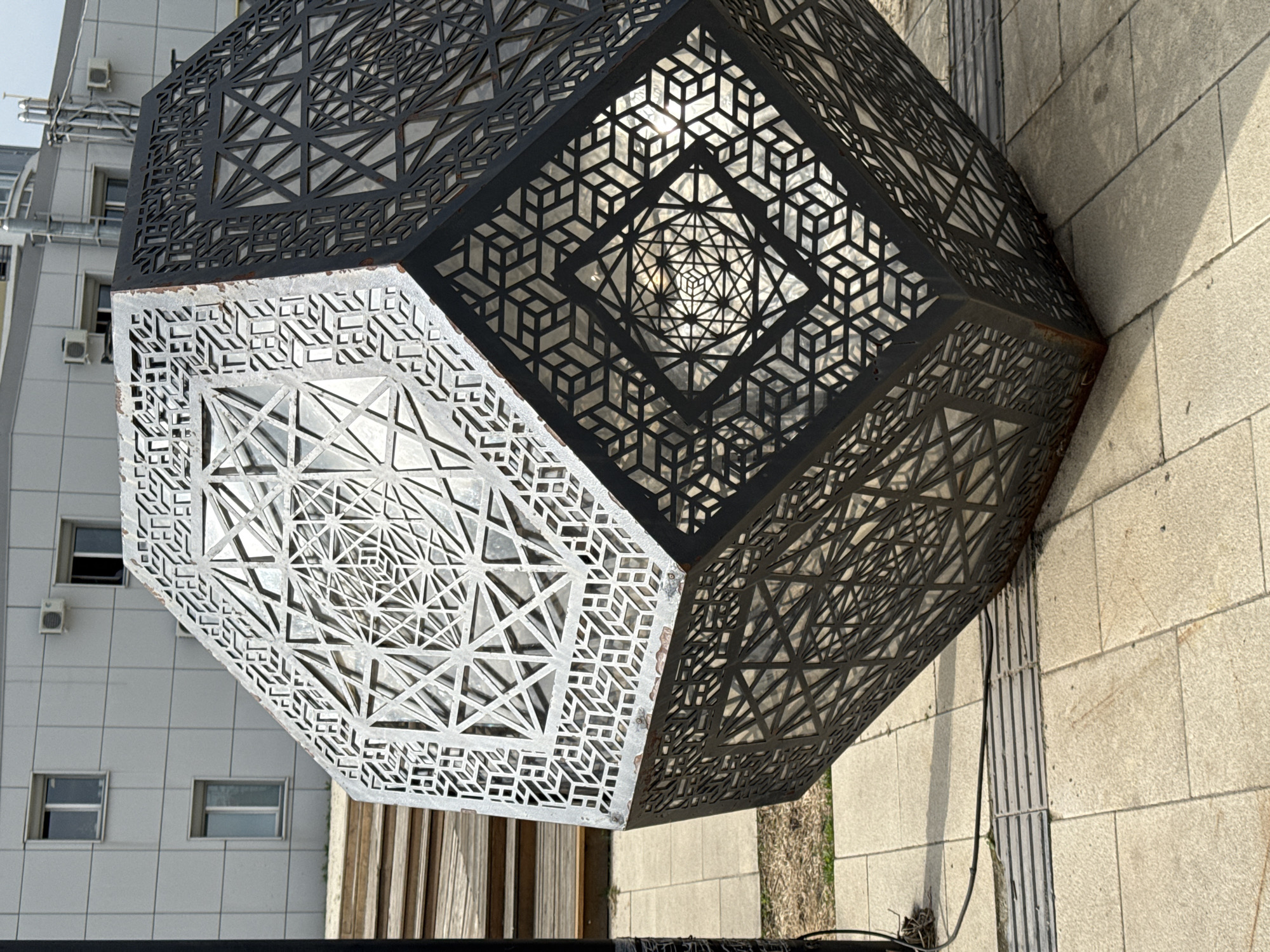 A metal sculpture with intricate geometric patterns, comprised of lighter and darker metal sections, rests on a paved surface in Nizhny Novgorod, Russia. The sculpture is a large, multi-sided polyhedron. The lighter-colored sections of the sculpture face a building with multiple windows and air conditioning units, while the darker sections align with the paved surface. A wooden structure is partially visible beneath the sculpture. A black cable runs along the paved surface near the sculpture.