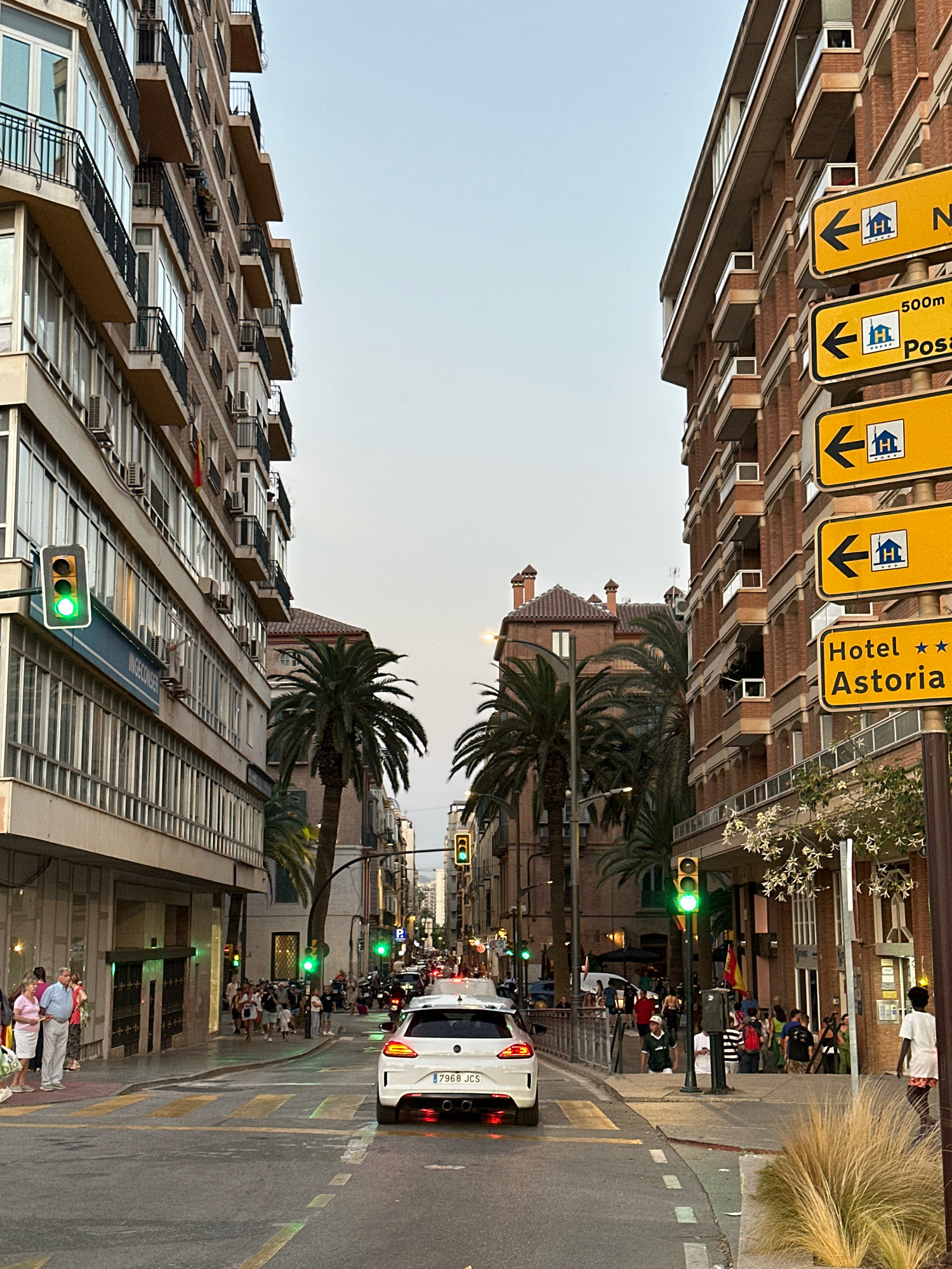 This is a street-level, medium-shot image captured in Málaga, Spain, likely in the late afternoon or early evening as indicated by the soft lighting. The scene is dominated by two multi-story brick apartment buildings flanking a city street. The building on the left features a modern facade with numerous balconies and large windows, while the building on the right has a more traditional brick exterior with similarly designed balconies and a street sign indicating "Hotel Astoria" with directional arrows and a distance of "500m".
The street itself is paved and marked with yellow and white lines, suggesting pedestrian crossings. Several palm trees are interspersed along the sidewalks, adding to the Mediterranean atmosphere. Traffic lights are visible, with one clearly showing a green light.
In the foreground, a white hatchback car with a Spanish license plate (7968 JCS) is positioned in the right lane. On the left sidewalk, a group of people, including two older individuals dressed in casual summer clothing, are standing near a building entrance. On the right sidewalk, a diverse crowd of pedestrians is walking and congregating. One person, wearing a white t-shirt and shorts, is walking away from the camera on the right edge of the image.
The sky is overcast and a pale gray. The overall impression is of a bustling urban environment during a pleasant, warm evening.