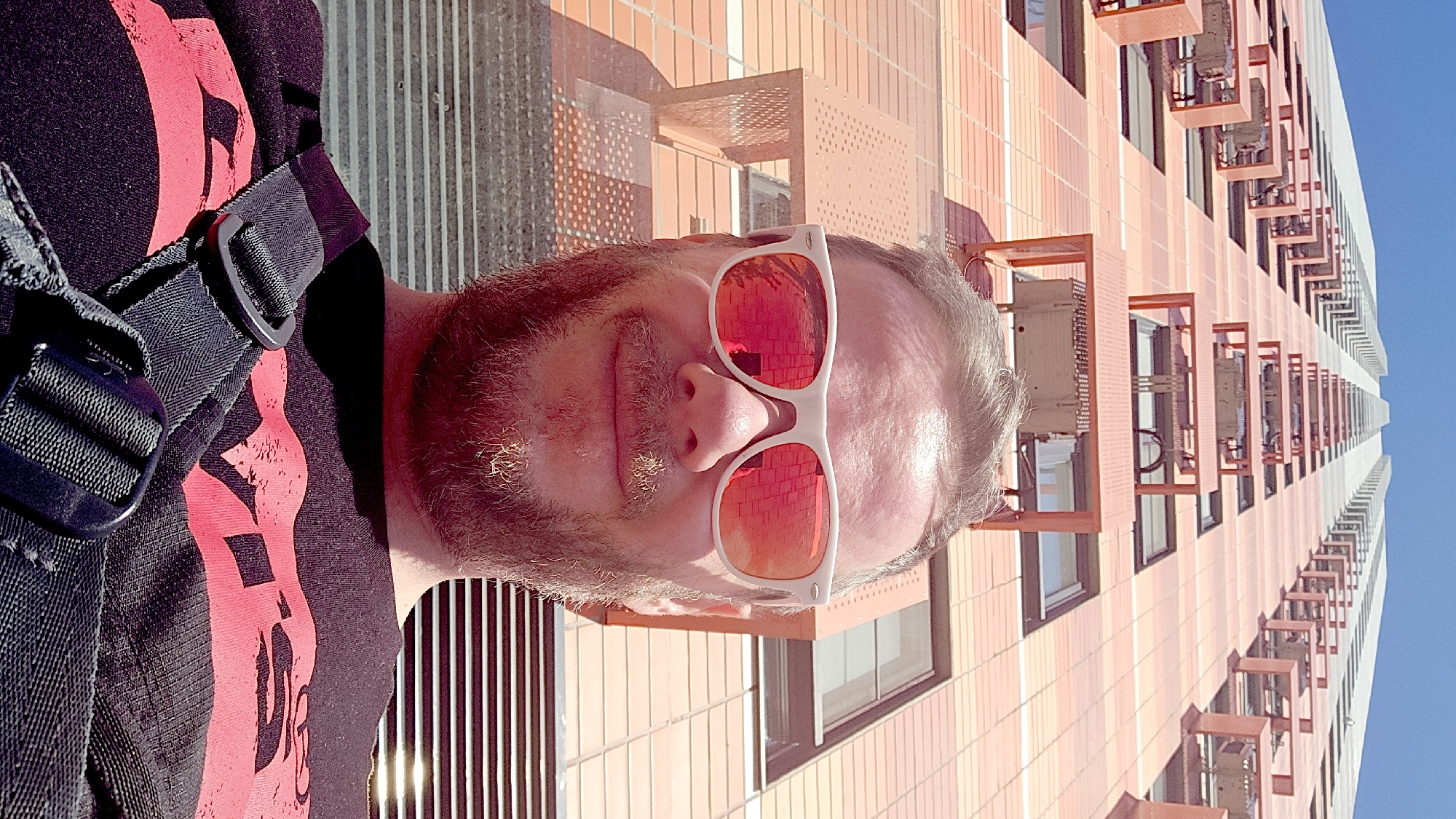 A selfie shows a man with a beard and sunglasses, standing in front of a modern building. The building is tall and has a patterned orange and white facade with projecting architectural elements. The man is wearing a black t-shirt with a large red design on it and a black strap crossing his shoulder. The location is Moscow, Russia.