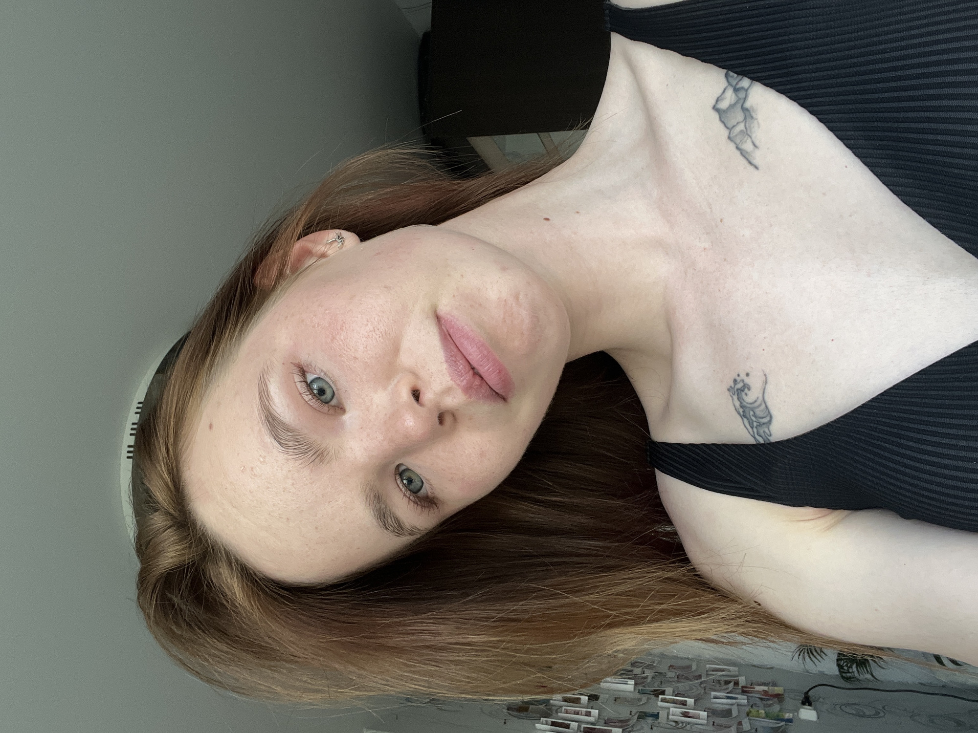 A close-up photograph shows a light-skinned woman with long, reddish-brown hair and light blue eyes. She is wearing a black tank top. She has two tattoos on her upper chest: one resembling mountains on her right and another on her left that appears to be of a whimsical, cartoon-like figure. The background includes a grey wall and some indiscernible objects. The picture is angled. There is no indication of the location in the city of Chaikovsky, Russia.