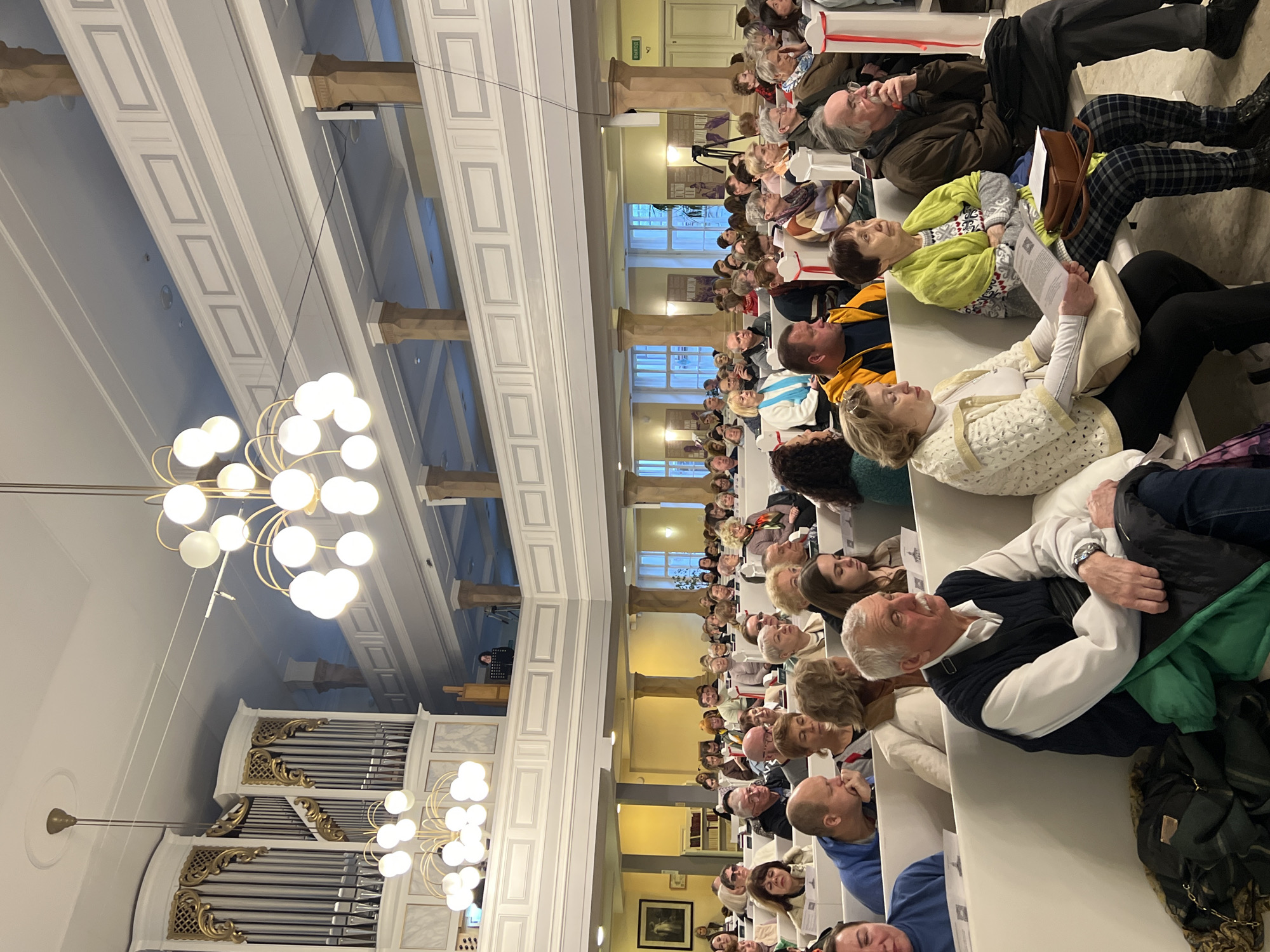 The photograph depicts the interior of a building, likely a church or concert hall, filled with a large crowd of seated people. The architecture features high ceilings with ornate detailing, including columns, coffered ceilings, and decorative moldings. A large pipe organ is visible on the left side of the image. There are multiple rows of white benches filled with attendees, primarily older adults. The people are sitting attentively, facing forward, suggesting they are watching a performance or listening to a speaker. A chandelier with spherical lights hangs from the ceiling. The image location is Saint Petersburg, Russia.