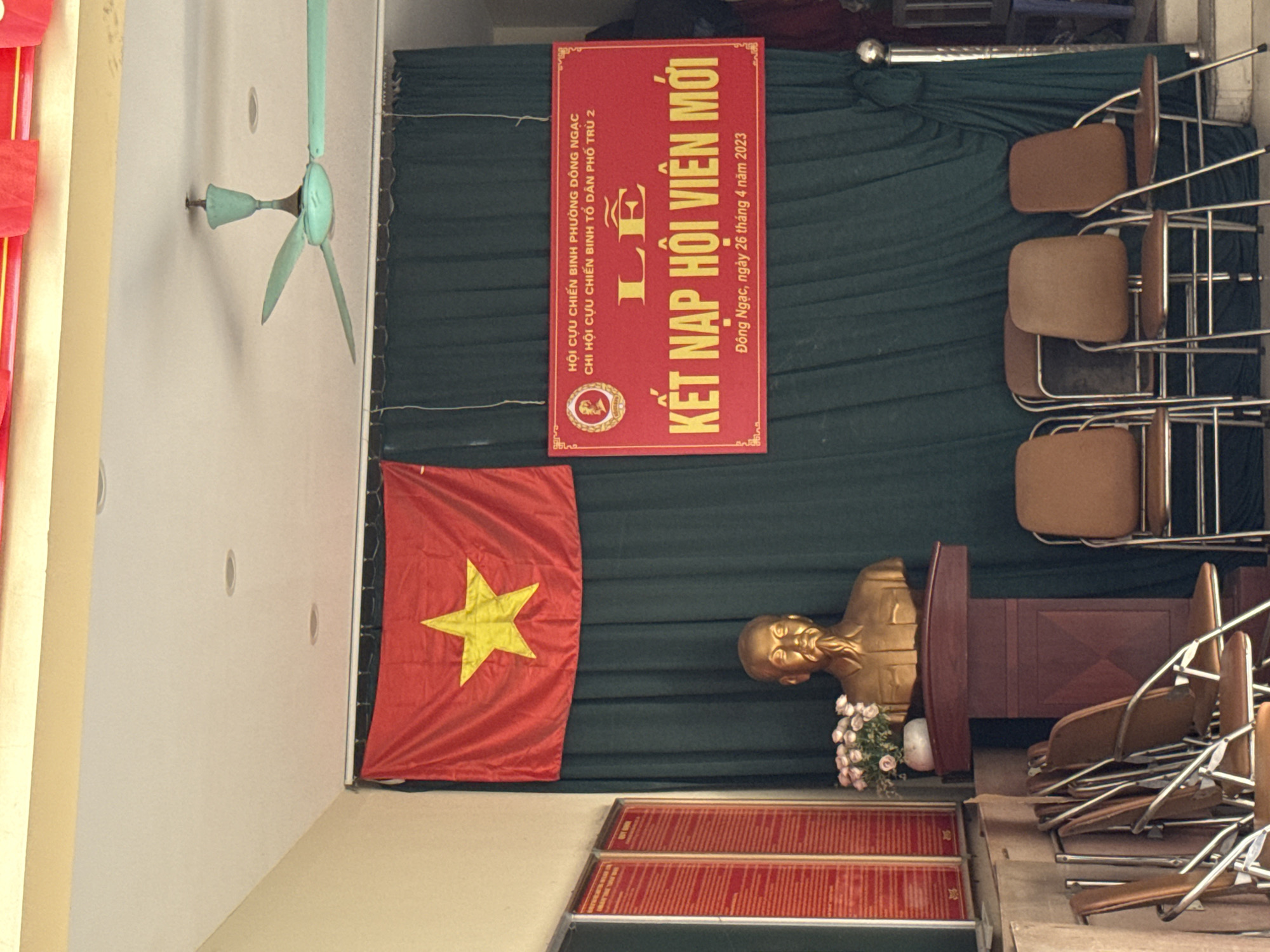 Here is a description of the image:
The image appears to be an indoor shot of a meeting or gathering space in Hà Nội, Vietnam. A green backdrop drapes behind the scene. A banner in Vietnamese, likely advertising an event related to "Hội Cựu Chiến Binh" (Veterans Association), is hung. The date on the banner indicates April 26, 2023. A Vietnamese flag hangs to the left of a golden statue of Ho Chi Minh, the revolutionary leader of Vietnam. A podium with flowers rests near the bust. A stack of chairs is on the right side. A ceiling fan is present.