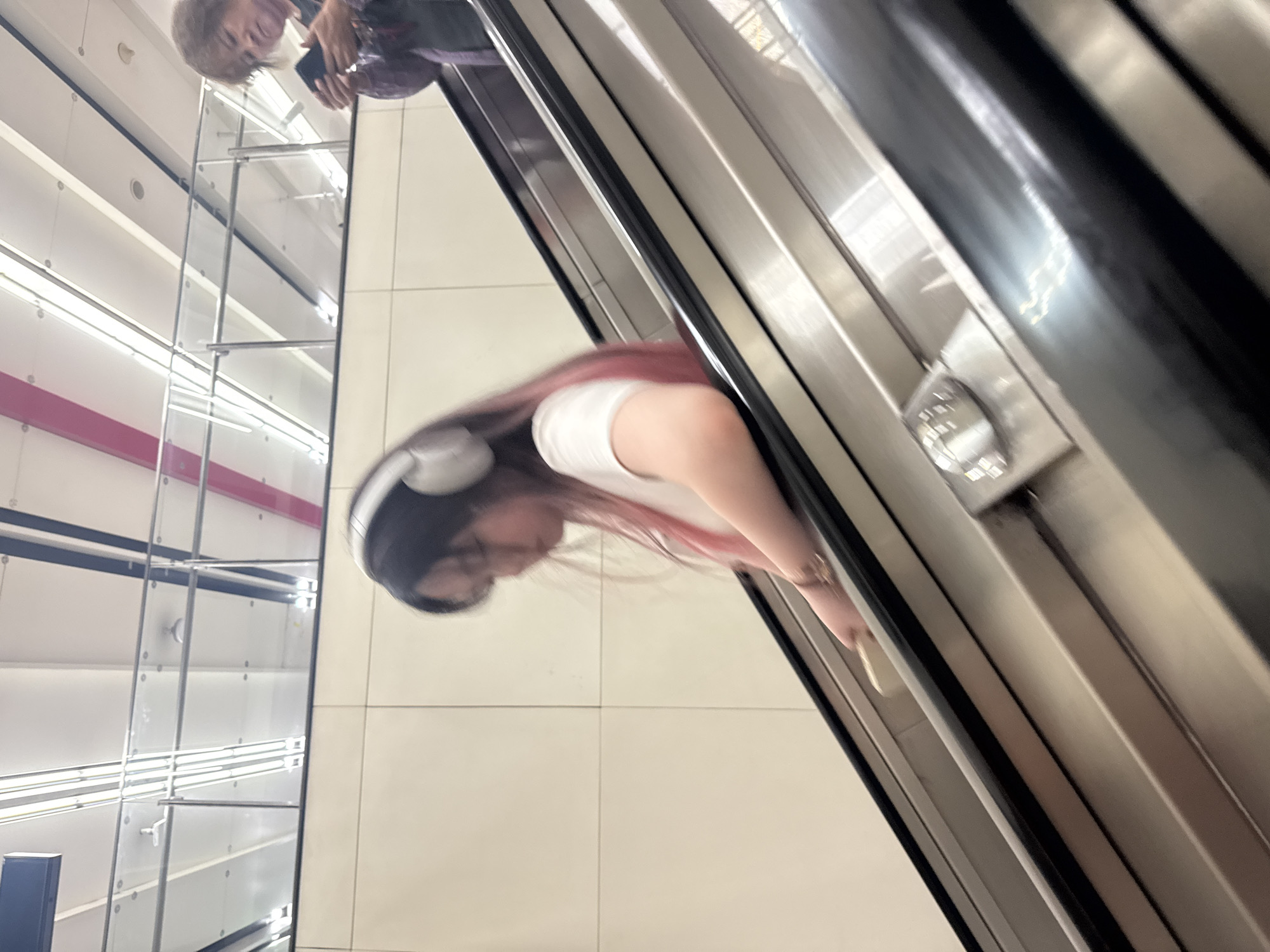 An outdoor escalator descends, with a young woman on the left and an older woman on the right. The young woman, with pink-streaked hair, wears headphones and a white t-shirt. She is in motion, appearing to look down and hold something light-colored. The older woman, in the upper right, is also in motion, looking at her phone. The environment is a clean, modern urban setting with a light-colored tiled wall, metallic escalator handrails, and bright overhead lighting. The background shows a railing and ceiling structure with fluorescent lights, suggesting a public transit station or similar indoor/outdoor transit area within Xicheng District, China.