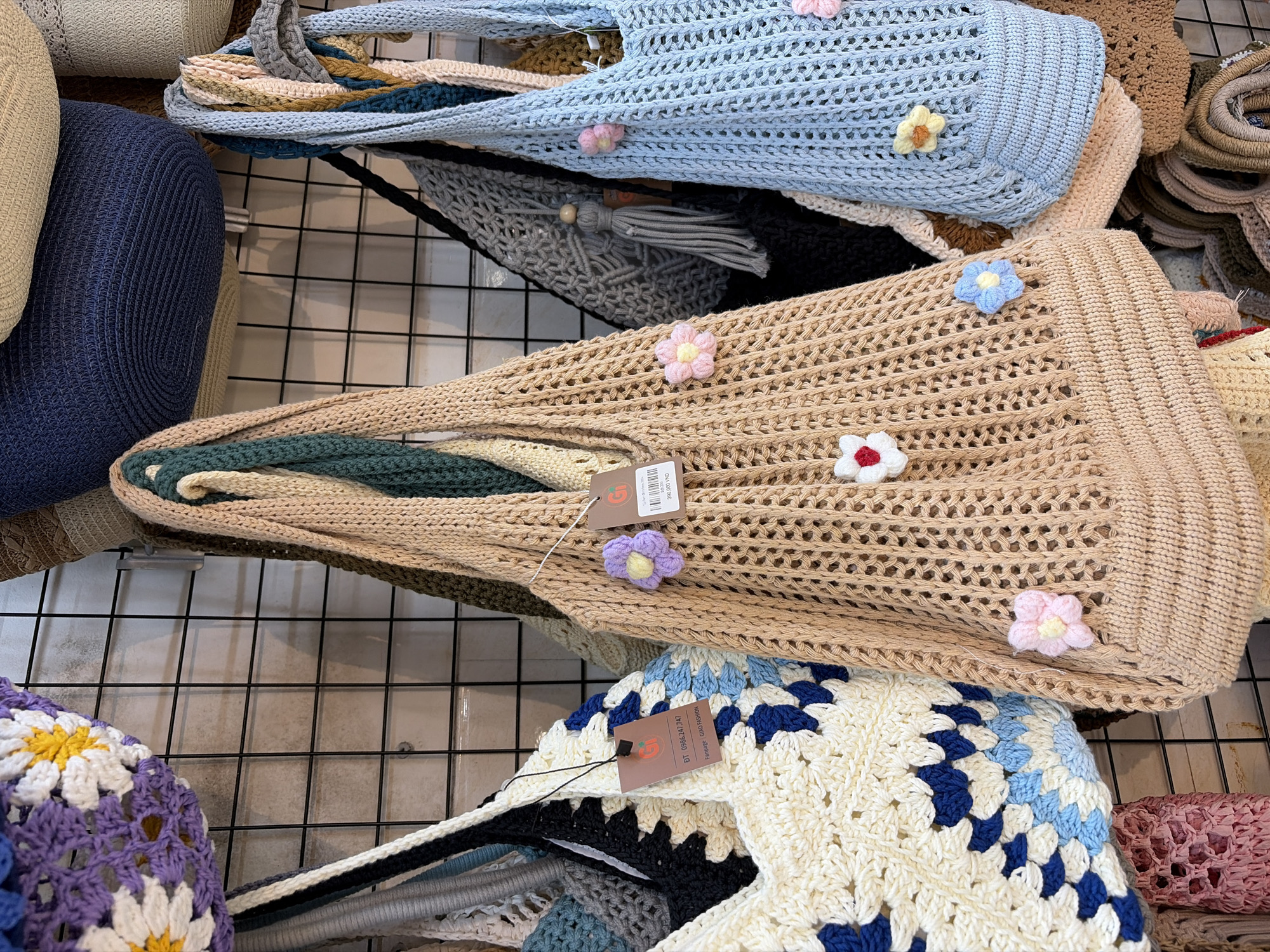 This is a medium shot, eye-level, indoor photograph of a display of crocheted bags at a market stall in Da Nang, Vietnam. The bags are hanging on a black wire grid against a light-colored wall. In the foreground, there are several crocheted tote bags with a net-like pattern and floral embellishments. From left to right, the first visible bag is cream-colored with a large blue and white granny square pattern. Next to it is a tan bag with a net pattern decorated with small crocheted flowers in pink, blue, and white. To the right of the tan bag is a light blue bag with a similar net pattern and a pink flower. Behind these, more bags are visible, including a darker tan bag, a gray bag with a tassel, and other multi-colored bags partially obscured. The lighting is soft and diffused, highlighting the textures of the crocheted items. The overall scene suggests a handicraft market or a small boutique selling handmade goods. There are no people visible in the frame.