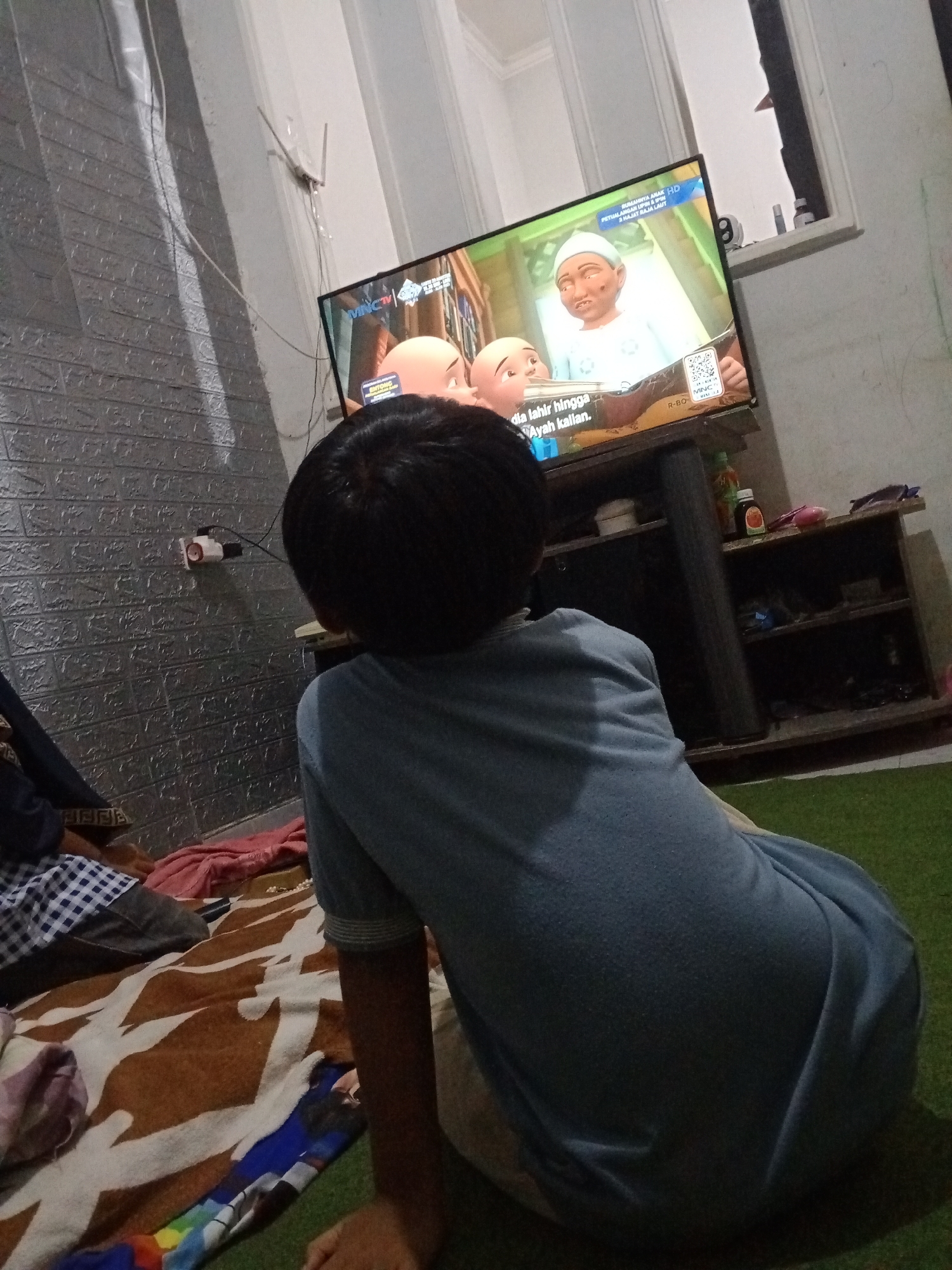 A young boy, seen from behind, sits on the floor facing a television screen in a room in Kemulan, Indonesia. The boy wears a light blue t-shirt. He is positioned on a green carpet, with a patterned blanket spread out in front of him. The television displays an animated scene with two characters, one wearing a white cap. Text in Indonesian is visible on the screen. Adjacent to the television is a dark wooden entertainment unit with shelves holding various items, including bottles. The wall behind the television is white, and to the left is a textured, grey wall. A small white object with wires is mounted on the grey wall. The lighting in the room appears to be artificial, casting shadows.