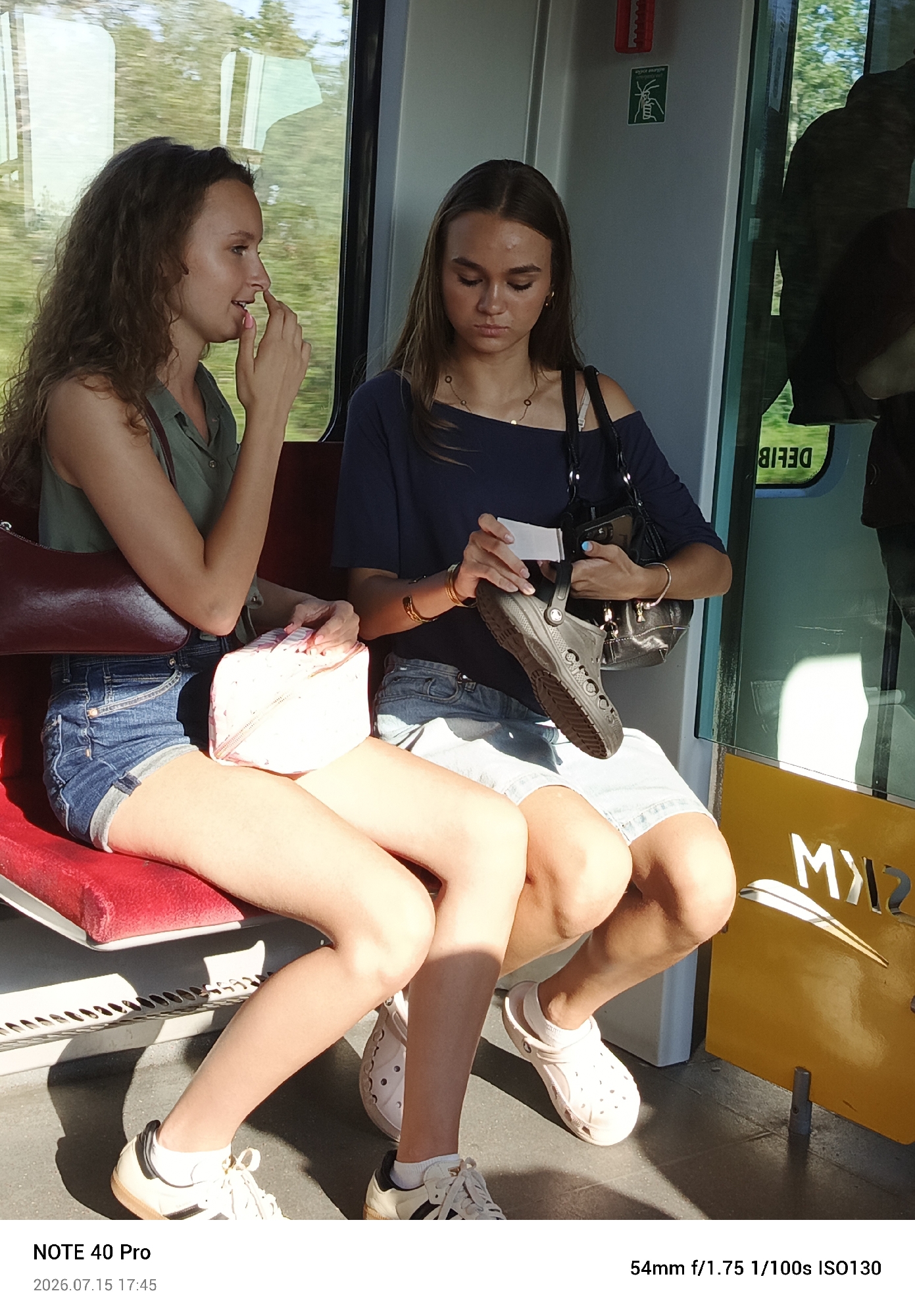 Two young women are seated on a red bench inside a train car. The woman on the left has long, curly brown hair and is wearing a green shirt and denim shorts. She is holding a dark red purse and a light pink makeup bag. The woman on the right has straight brown hair and is wearing a dark blue off-the-shoulder top and denim shorts. She is holding a black purse and a black Croc shoe. Both women appear to be looking down. The train car interior is visible, with a window showing trees and greenery outside. Text on a yellow panel reads "MKS". The timestamp on the image indicates it was taken on July 15, 2026, at 17:45.