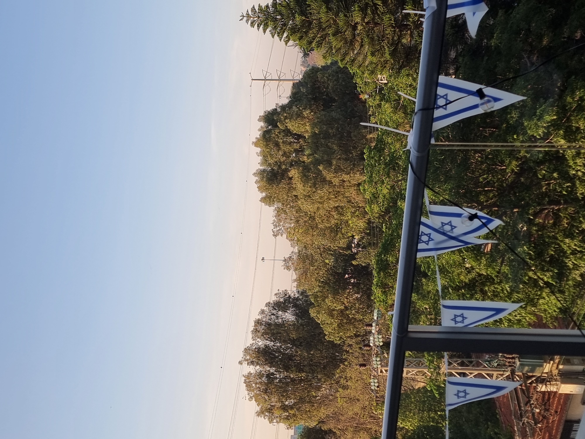 The image shows a vertical outdoor scene. In the foreground, a vertical metal pole holds a string of triangular Israeli flags, interspersed with small light bulbs. The background features dense green trees under a light blue sky. Power lines are visible through the trees. The location is Ramat Gan, Israel.