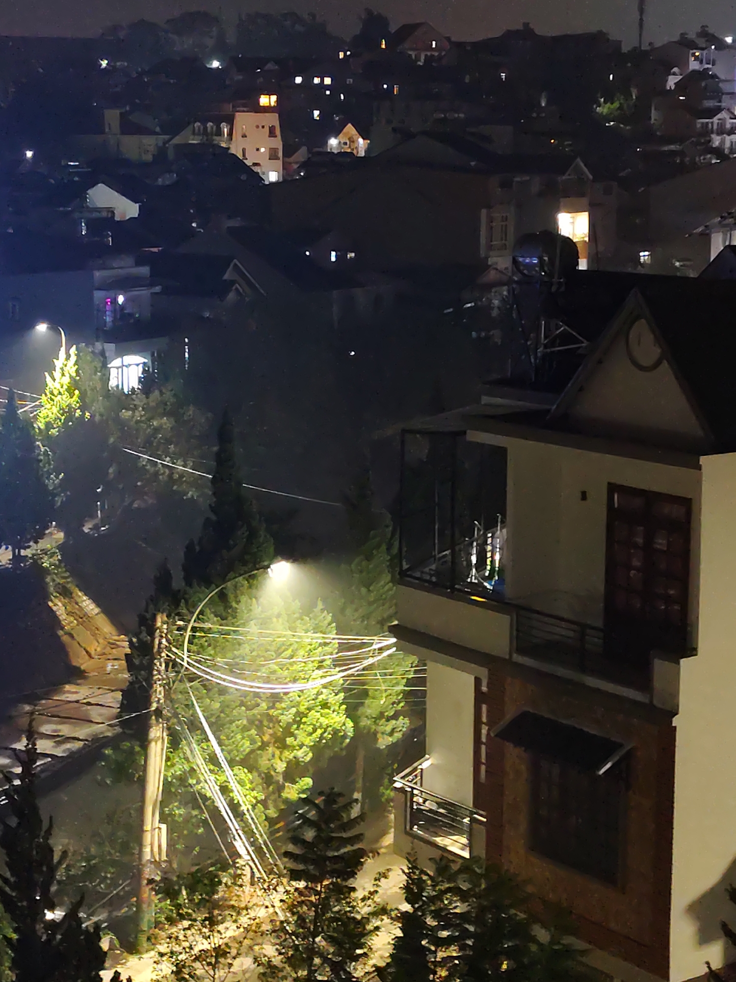 The image depicts a night view in Da Lat, Vietnam. The foreground shows a multistory building with balconies, one enclosed with glass. To the left, there's a street scene illuminated by a streetlight, with power lines and trees obscuring the view. In the background, there are other buildings scattered across a hillside, with various lights indicating residential activity. The overall scene has a hazy, damp quality, typical of Da Lat's climate.