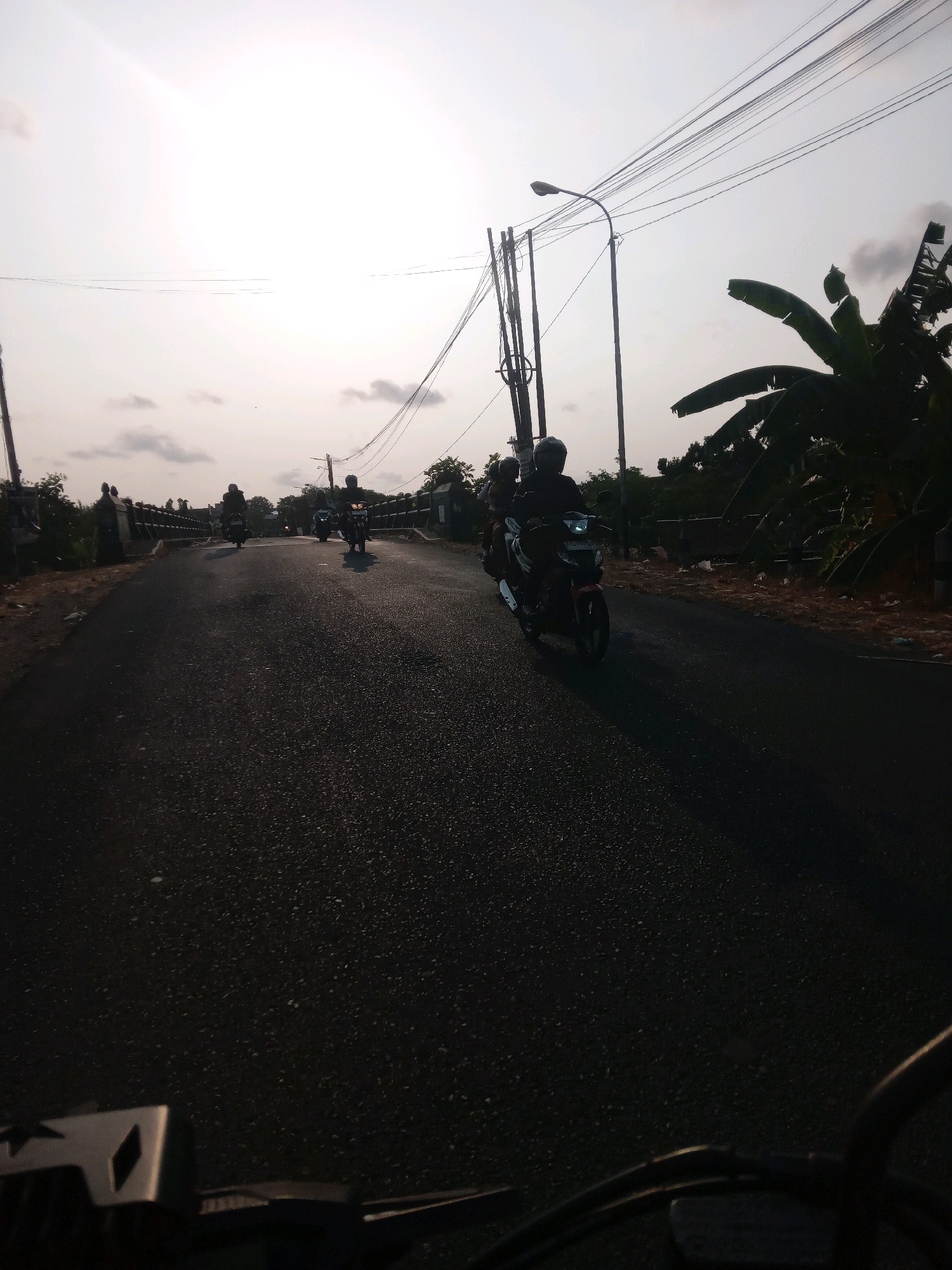 The image depicts a road in Banguntapan, Indonesia, with several motorcycles driving on it. The sun is low in the sky, casting long shadows and creating a bright, hazy atmosphere. A street light is visible on the right side of the road, along with telephone poles and wires. There are trees and vegetation along the sides of the road, and a bridge is visible in the background. The motorcycles appear to be moving in the same direction, with some riders carrying passengers. The overall scene suggests a typical day in Banguntapan, with the road being used for daily transportation.