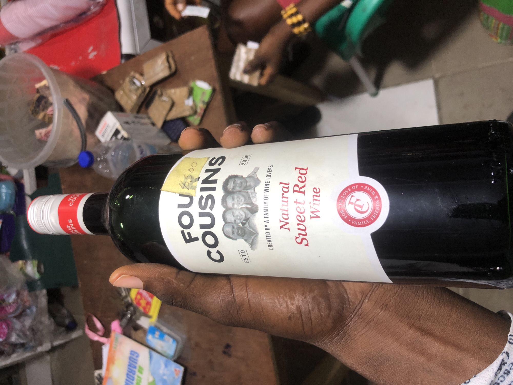 A person's hand holds a bottle of "Four Cousins Natural Sweet Red Wine" in what appears to be a market or shop. The bottle has a white label with the brand name in large black letters, followed by an illustration of four men's faces. Below this, it states "CREATED BY A FAMILY OF WINE LOVERS." The wine is described as "Natural Sweet Red Wine." A circular red seal with the initials "FC" and text "FOR THE LOVE OF FAMILY, FRIENDS & WINE" is visible at the bottom of the label. In the background, various items are visible, including plastic containers, bags, and other products, suggesting a retail environment. The presence of a person's hand suggests the item is being handled or presented. The lighting is adequate, and the image is in focus, allowing for clear identification of the product. The location is indicated as Ado, Nigeria.