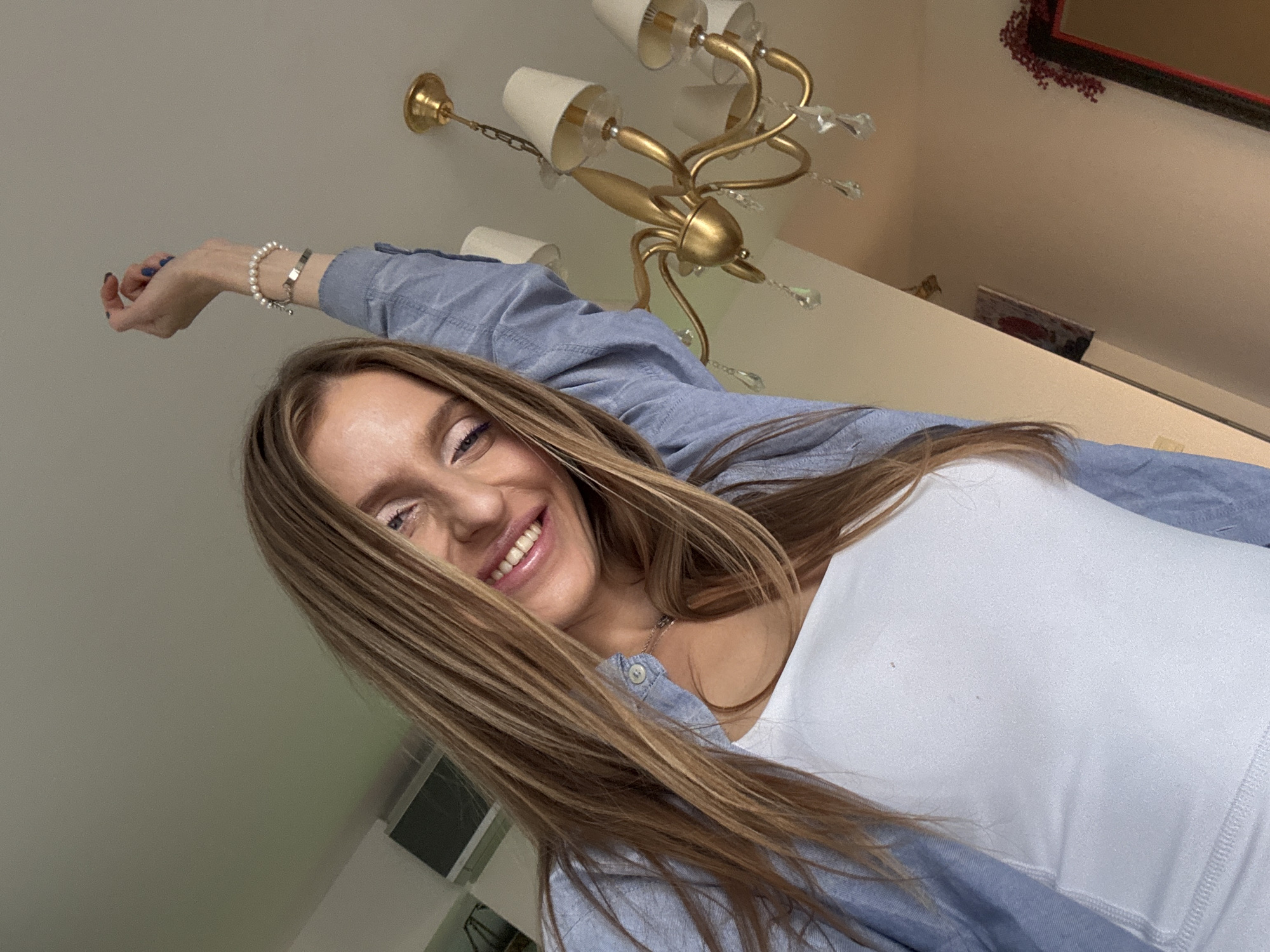 The image captures a low-angle shot of a young woman indoors. She has long, light brown hair that falls over her shoulders and a bright smile. She is wearing a white tank top and a light blue unbuttoned shirt over it. Her right arm is raised above her head, and she is wearing a pearl bracelet and a silver bracelet on her wrist. The background features a golden chandelier with crystal pendants hanging from it and a beige-colored wall. To the left, a portion of a dark-framed mirror with red decorations is visible. A dark rectangular object, possibly a framed picture or a screen, is also present on the wall. The lighting appears to be natural, casting soft shadows. No specific details of Budva, Montenegro are discernible from the image.