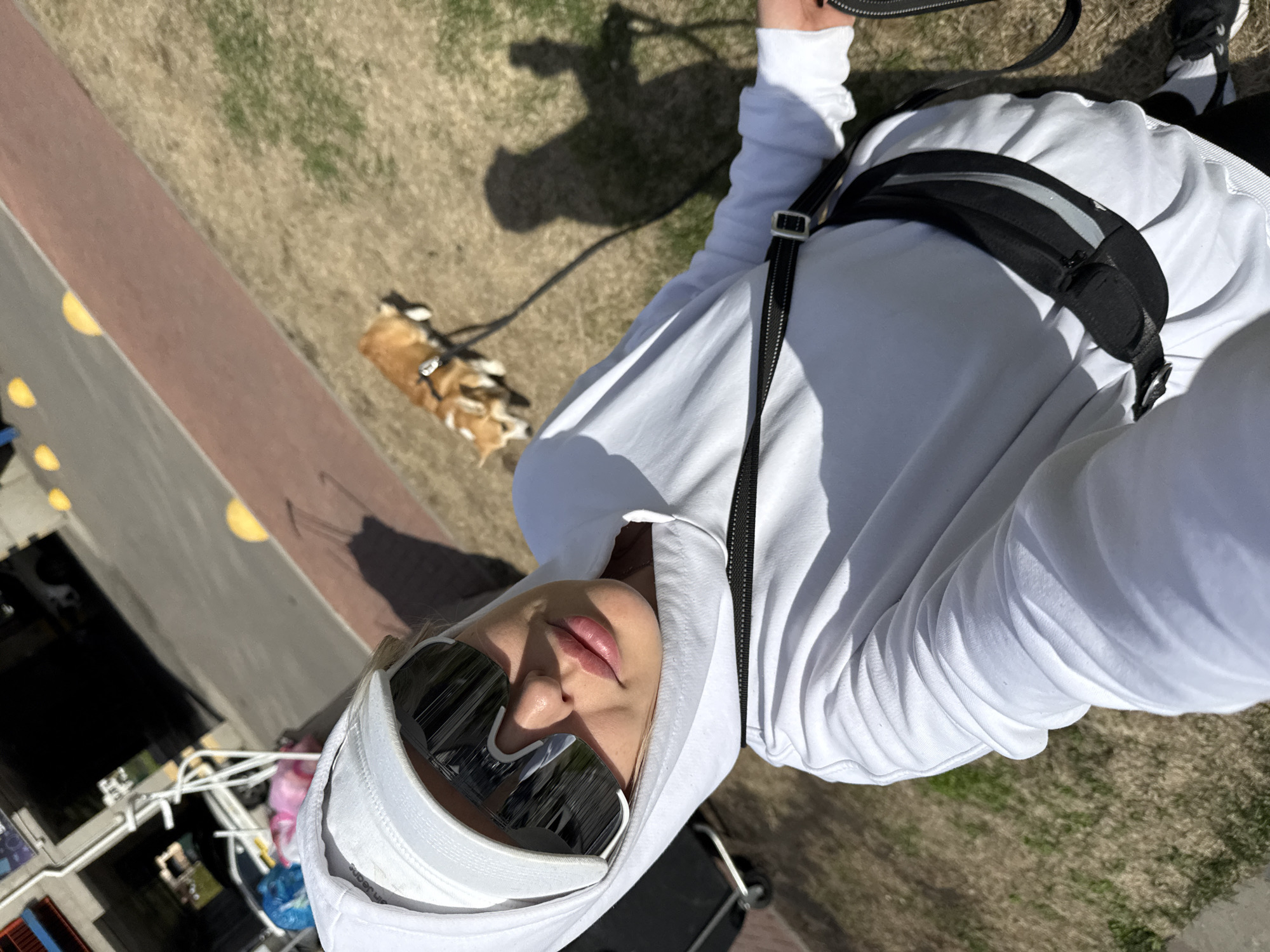 A selfie, taken in daylight in Krasnoyarsk, Russia, depicts a person wearing a white hoodie, a white baseball cap, and mirrored sunglasses, looking down at the camera. They have on a black belt bag and a black strap crossing their chest. A small dog is on a leash near a grassy area. The background shows a walkway and structures, potentially part of a park or outdoor setting.
