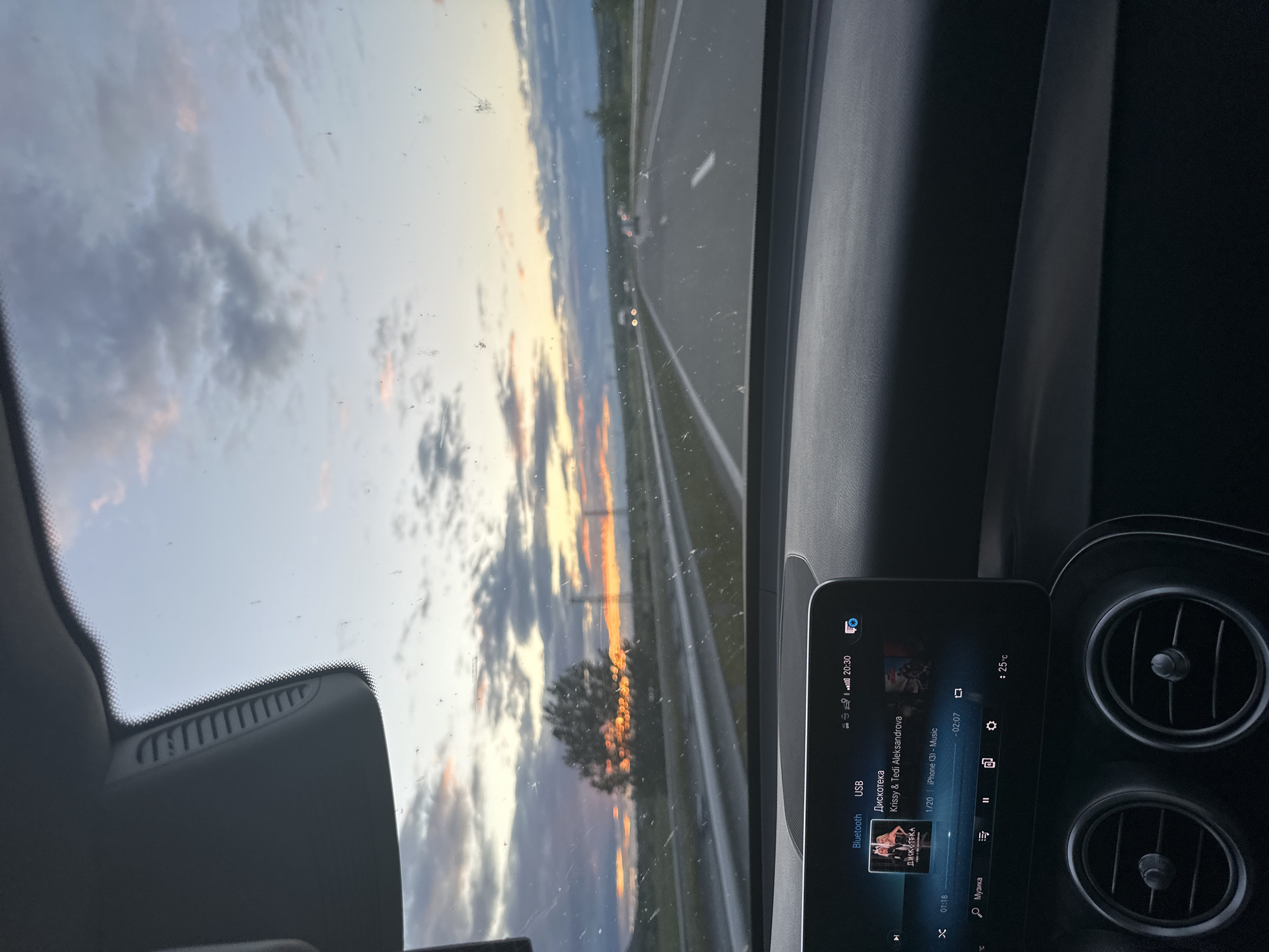 The image captures an interior view from a vehicle, likely a car, showcasing a sunset seen through the windshield. The sky displays a gradient of colors with clouds tinted with shades of orange and blue. An asphalt road stretches into the distance. The dashboard of the vehicle is visible, including a display screen set to play music with the song "Дискотека" by Krissy & Tedi Aleksandrova selected from the iPhone (3) USB music source with timestamp 20:30 and inside temperature +25°C. Based on the song title, the location might possibly be in the city of Krum, Bulgaria.