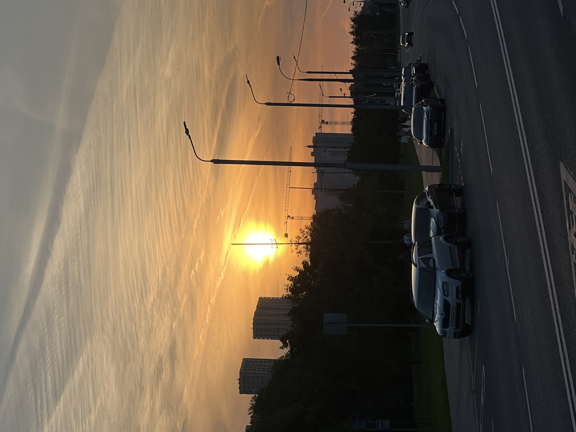 This image depicts an urban street scene during either sunrise or sunset in Moscow, Russia. The sky occupies the upper half of the frame, transitioning from a brilliant orange and yellow glow at the horizon to lighter peach and greyish-blue tones overhead, interspersed with faint, wispy cloud patterns and distinct contrails. The sun, a bright disk centered in the left mid-ground, casts strong backlighting across the scene.
On the left side, a dense line of dark-foliaged trees and bushes runs parallel to the road. Behind these trees, two light-colored, multi-story buildings, likely residential or office towers, are visible, partially silhouetted against the bright sky. A white rectangular sign stands in front of the trees. Dark silhouettes of multiple individuals are discernable walking on a path behind the trees.
The asphalt road in the foreground features prominent white double solid lines marking the center and a single white solid line delineating the right shoulder. A white Skoda Yeti SUV, bearing the Russian license plate "P317OM 177" (the "177" region code indicating Moscow), is parked on the left side of the road. Behind it, a dark-colored sedan is also parked.
On the right side of the road, a series of tall streetlights with curved arms and horizontal fixtures extends into the distance, their forms largely silhouetted. Beyond them, several tall construction cranes and the upper portions of a large building under construction are visible, indicating ongoing development. Further down the road on the right, additional individuals are seen as dark silhouettes, walking on a sidewalk. Various other vehicles are parked or driving along the curving road, which recedes into the background.