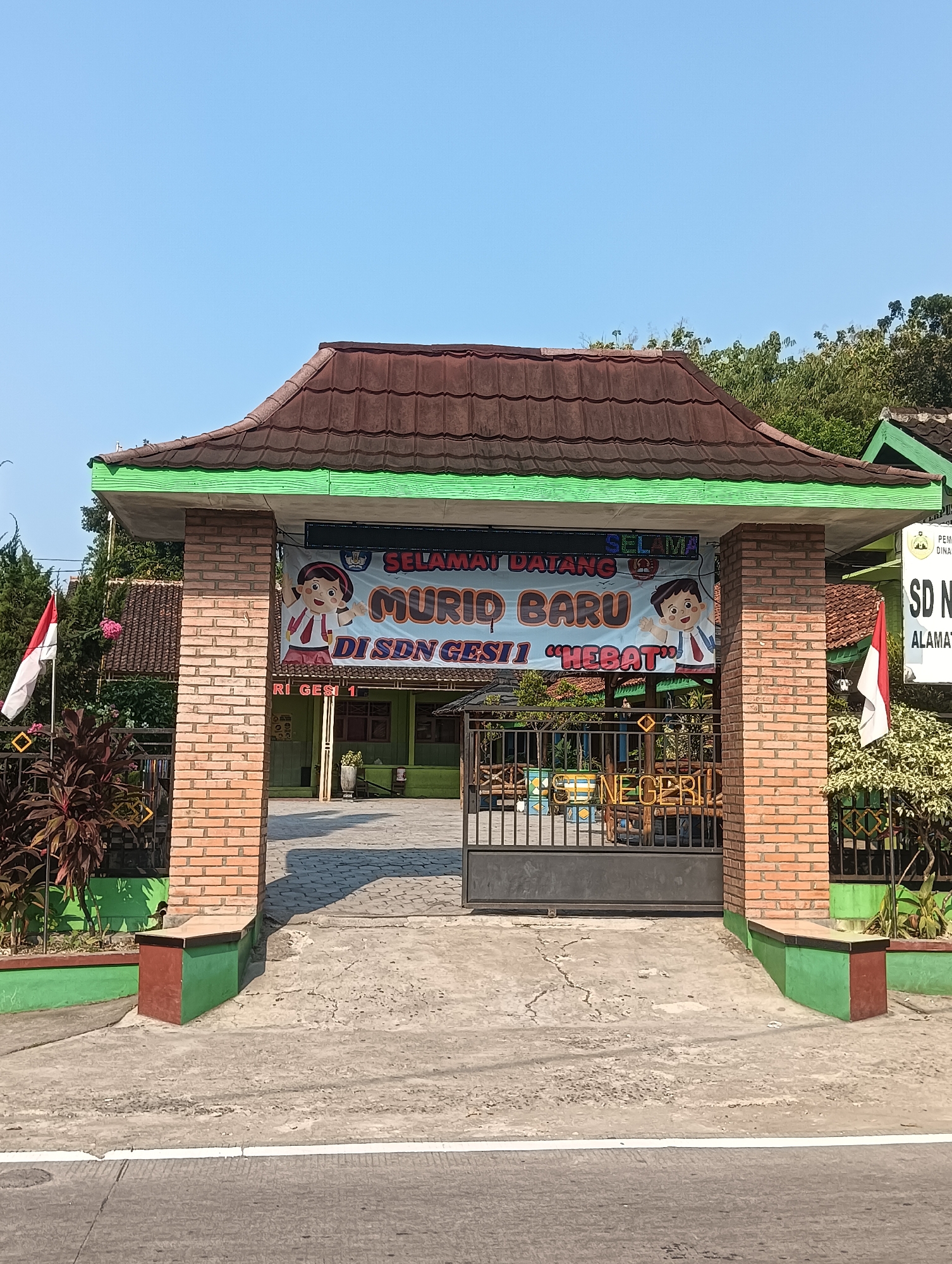 The image shows the entrance to SDN GESI 1, a public elementary school located in Palemrejo, Indonesia. The entrance features a brick gate with a tiled roof. A banner above the gate welcomes "MURID BARU DI SDN GESI 1 'HEBAT'", which translates to "New Students at SDN GESI 1 'Great'". The school grounds are visible beyond the gate, with a green building and trees. A flag of Indonesia is planted to the left of the entrance. The scene is bathed in bright sunlight under a clear blue sky.