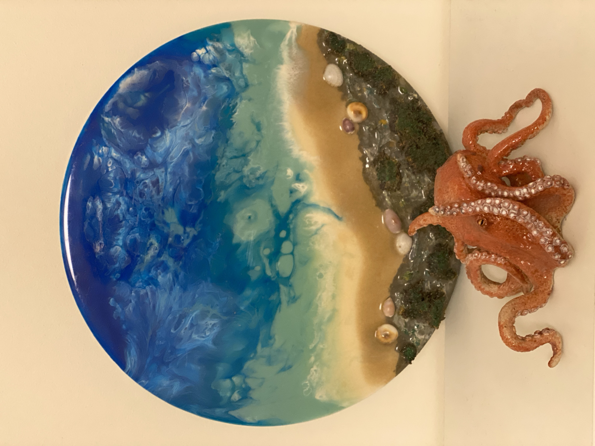 This is a studio photograph of a circular resin art piece depicting a stylized ocean scene with a miniature octopus figure. The art piece is divided into three distinct sections. The largest section is a swirling depiction of the ocean, rendered in shades of deep blue, turquoise, and white, with white accents mimicking sea foam and waves. To the right of the ocean is a sandy beach, depicted in beige and yellow tones, with a thin line of white representing the surf. The rightmost section represents a rocky shoreline or seabed, with dark green moss-like textures and small, smooth stones in various light colors embedded within the resin. An orange, three-dimensional octopus figure is placed on the right edge of the art piece, with its tentacles extending outward and partially over the sandy beach section. The octopus appears to be reaching towards the water. The overall scene is a miniature, idealized representation of a coastal environment. The lighting is even and diffused, suggesting an indoor studio setting, and there is no indication of time of day or weather conditions. No visible text is present in the image. The location context of Moscow, Russia, has no bearing on the visual content of the image.