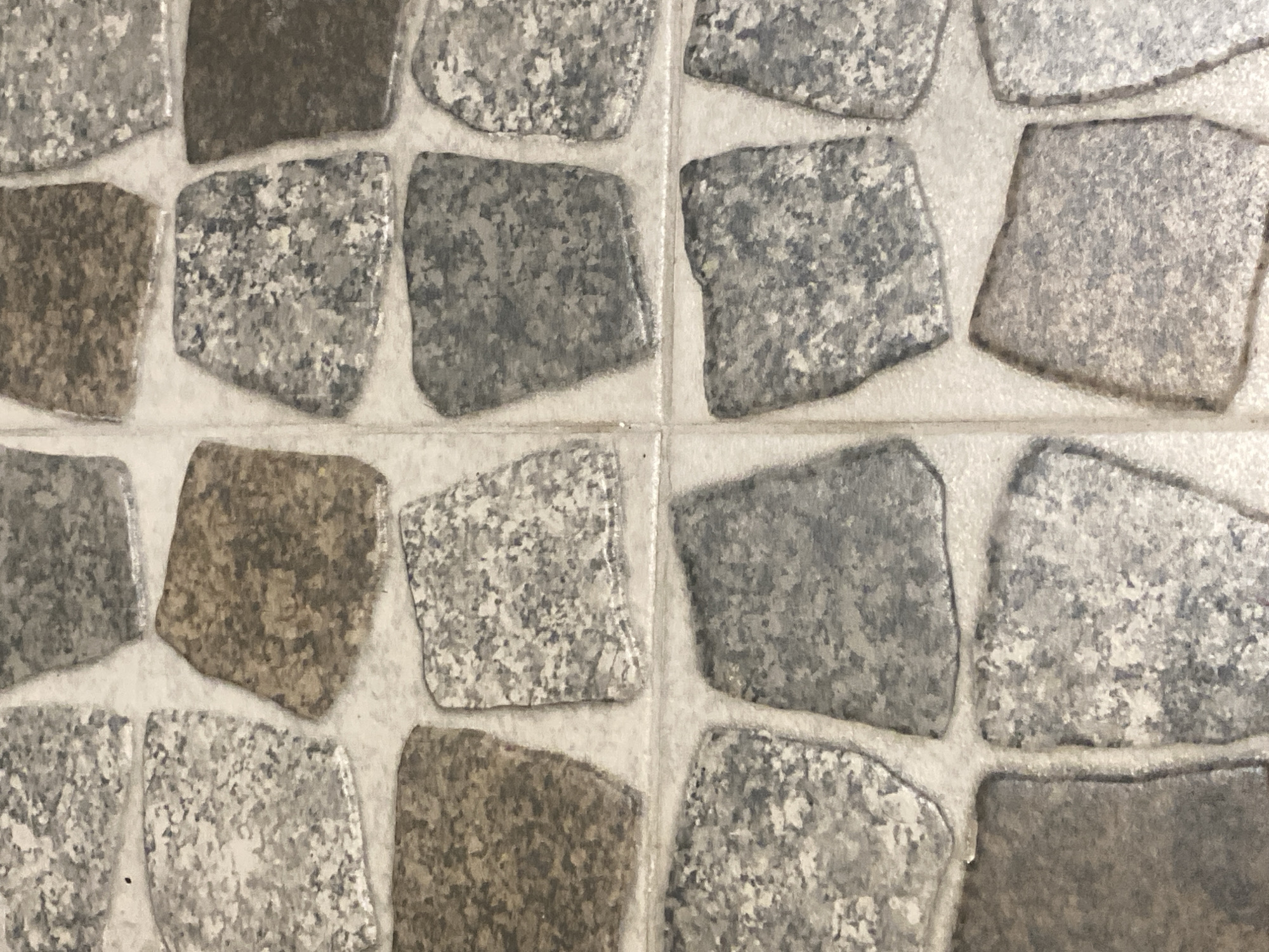 The image displays a close-up, overhead view of a tiled surface. The tiles are irregularly shaped and arranged in a staggered pattern, resembling cobblestones or natural stone flagstones. They vary in color, with shades of gray, white, brown, and speckled patterns reminiscent of granite. The grout between the tiles is a light, off-white color. The lighting appears even, with no strong shadows, suggesting an indoor environment or an overcast day. The image is devoid of any human presence or other identifiable objects or actions. There is no information within the image or its context to suggest a specific location within Chișinău, Moldova.