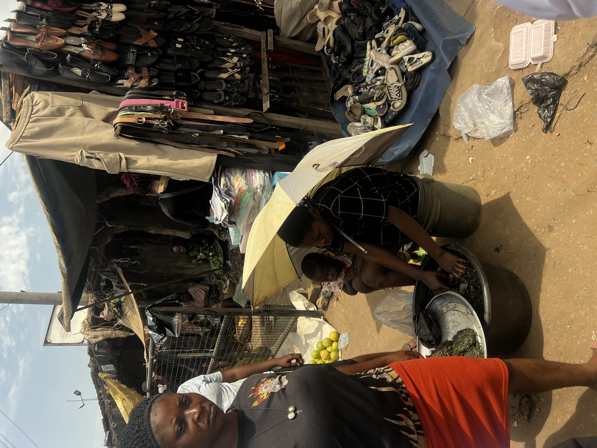 The image shows a street scene in Unknown, Unknown. A person in an orange skirt lies horizontally at the bottom of the frame. A second person stands under a white umbrella washing greens in a tub, possibly with a child present. Merchandise, including shoes, belts, and clothing, is displayed in stalls or on the ground. Lemons are on the ground near the person in the orange skirt. Additional individuals are visible in the background. The location appears to be a bustling outdoor market or commercial area.