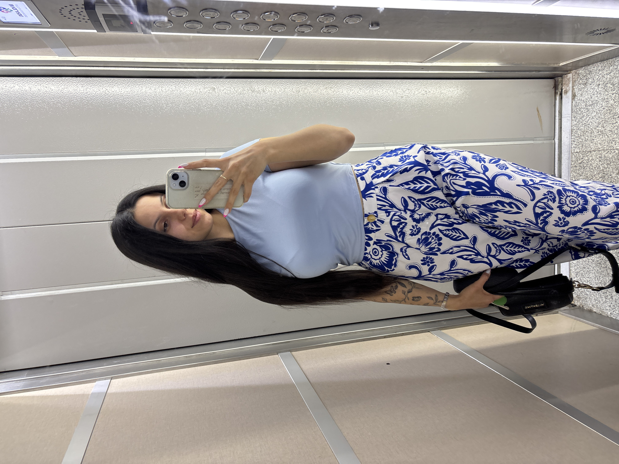A woman is taking a selfie in an elevator. She has long, black hair and is wearing a light blue shirt and blue and white patterned pants. She is holding a black purse. The elevator has metal walls and a control panel at the top. The location is likely in the city of Sliven, Bulgaria, but this is not explicitly verifiable from the image.