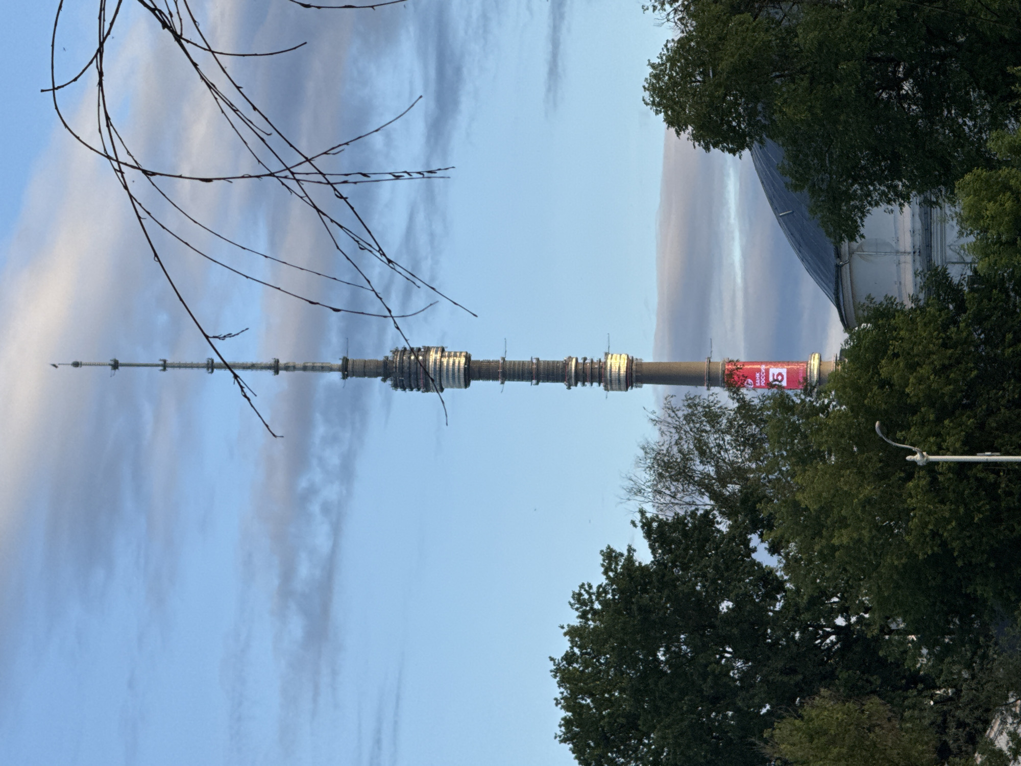 This media file captures a view of the Ostankino Tower in Moscow, Russia. The image is oriented vertically, presenting the tower as a prominent vertical element against a sky filled with wispy clouds. The time of day appears to be late afternoon or early evening, suggested by the soft, diffused lighting. The weather seems mild, with the sky a pale blue and the clouds indicating a possibility of light changes.
The subjects include the towering structure of the Ostankino Tower, a man-made object of significant height. Trees with dense green foliage frame the lower portion of the image and also appear on the right side, suggesting the tower is situated in a park-like or green urban area. Bare tree branches extend into the upper left quadrant of the frame, adding a natural, somewhat stark element to the composition.
A notable detail on the tower is a large, red banner with white text. While partially obscured and potentially blurry due to the image orientation and resolution, some of the text appears to be in Cyrillic. The visible characters suggest it could be a Russian phrase or advertisement, possibly related to an event or organization. The number "15" is clearly visible on the banner.
The scene is one of a prominent landmark set within a natural and urban environment. The overall impression is calm, with the vastness of the sky and the impressive height of the tower being key visual elements. There are no people explicitly visible in the frame, focusing the attention on the architecture and the surrounding environment. The lighting and the presence of lush trees suggest a pleasant day, potentially during warmer months.
