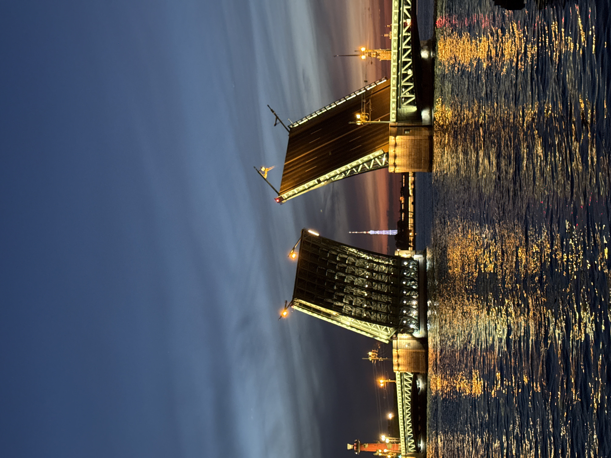 The image depicts the Palace Bridge (Dvortsovy Most) over the Neva River in Saint Petersburg, Russia, at night. The bridge's two central bascule leaves are fully raised, creating a prominent V-shaped opening for maritime traffic. The undersides of the raised bridge sections are brightly illuminated with numerous warm, yellow-orange lights, highlighting their structural trusses. The fixed bridge spans on both the left and right sides are also lit, featuring a distinctive green-painted lattice framework.
On the far left, a red Rostral Column stands illuminated with orange light. In the distant background, centered between the raised bridge sections, the Saint Petersburg TV Tower is visible, glowing with a faint purple light. Additional city lights and indistinct building silhouettes line the horizon.
The sky transitions from a deep, dark blue at the top to a faint orange and red hue near the horizon on the right, indicating either twilight or a lingering sunset. The Neva River occupies the lower half of the image, appearing dark blue with significant reflections of the bridge's yellow-orange lights, creating shimmering, elongated streaks across its rippled surface. No discernible individuals or vehicles are visible on the bridge or immediate foreground.