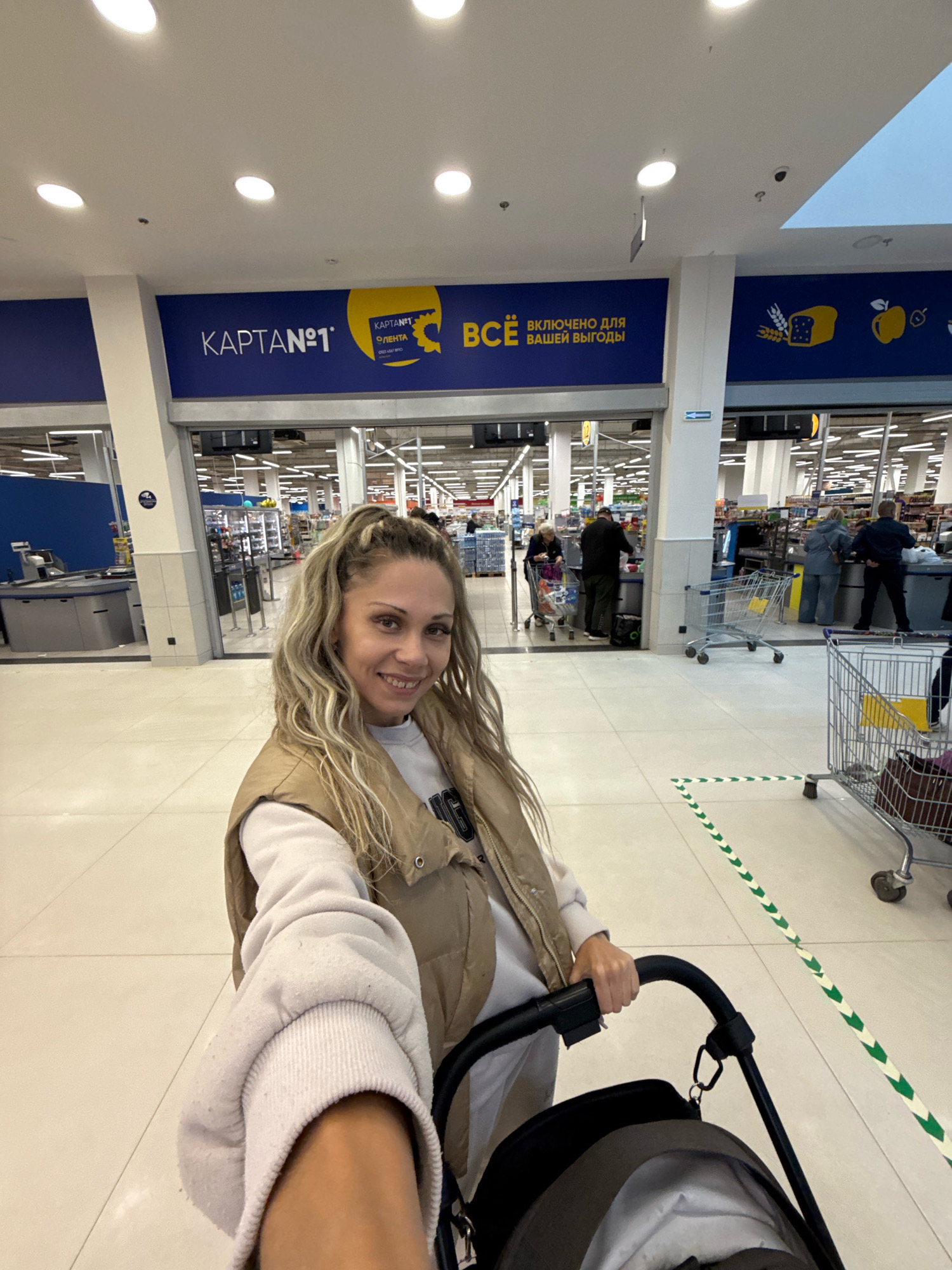 A woman is taking a selfie in a brightly lit supermarket in Bor, Russia. She is pushing a stroller and smiling at the camera. In the background, several people are visible at the checkout counters, and shopping carts are lined up. Signs in Russian advertising "Karta№1" and "VSE VKLYUCHENO DLYA VASHEY VYGO-DY" (everything included for your benefit) are displayed prominently on the wall. The floor is tiled with light-colored squares, and the ceiling features recessed lighting.