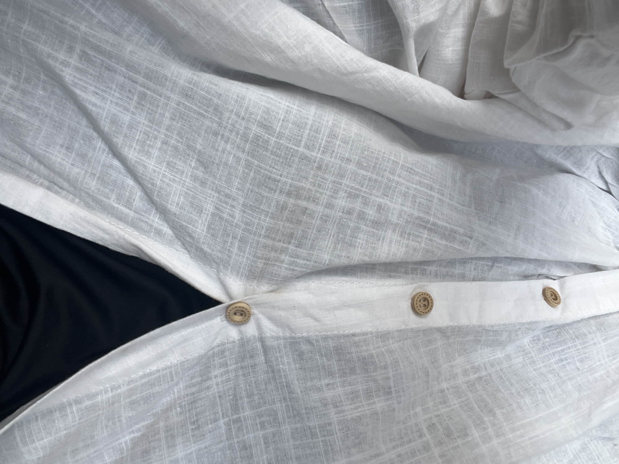 The image displays a close-up view of the top portion of a white, textured button-up shirt, likely made of linen or a similar lightweight fabric. The shirt is layered over a black garment, visible in the V-neck opening. Three wooden buttons with a carved smiley face design are visible on the placket. The fabric is creased and draped, suggesting it is being worn or has been recently handled. The image is a tight shot, focusing solely on the clothing item and its details, with no environmental context provided, and therefore no specific location in Teluk Air, Indonesia, can be determined.
