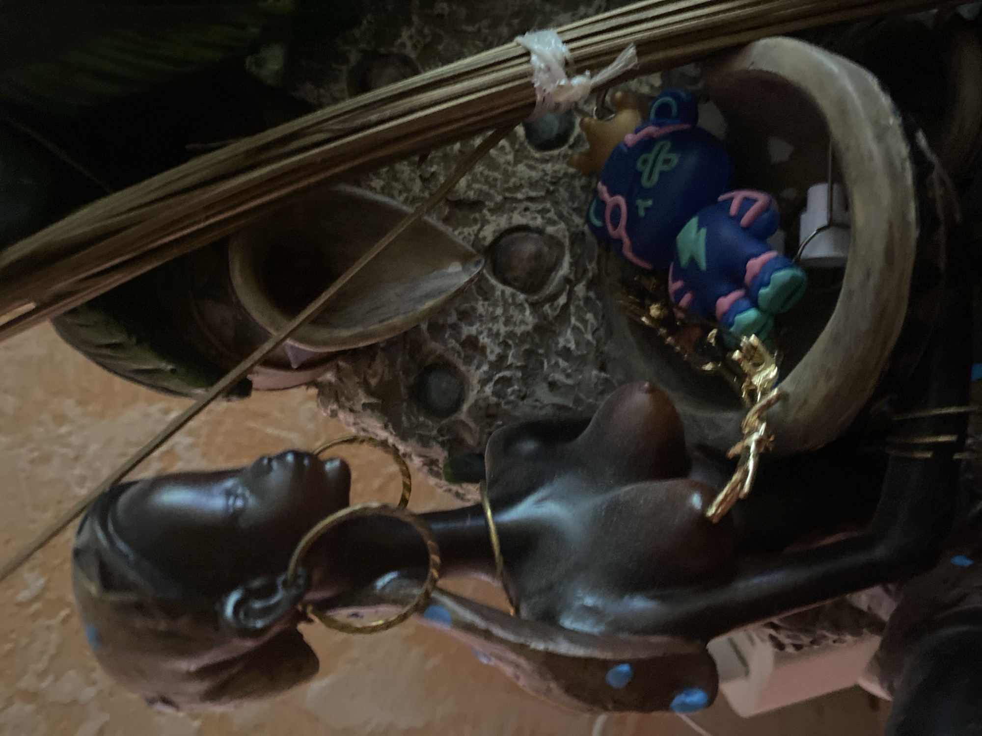 The image displays a close-up, low-angle view of a collection of objects, possibly part of a decorative arrangement or altar. The primary subjects are two dark-skinned figurines, one appearing to be a torso and bust with prominent breasts and jewelry, and the other a head and neck with large hoop earrings. The figurines have a smooth, dark finish.
Interspersed with the figurines are elements that suggest a natural or rustic theme. A bundle of thin, dry, straw-like stalks, possibly from a broom or grass, is prominently positioned in the upper half of the frame, tied with a white plastic strip. Behind this, a textured, rough surface, possibly stone or aged plaster, forms the background, with some circular indentations.
Also visible is a cartoonish, blue and pink figurine with large eyes and a stylized, almost alien-like appearance, positioned to the right of the straw bundle. It features geometric patterns and bright colors. A curved, light-colored object, resembling a horn or a large shell, arches over the cartoonish figurine. A gold-colored chain or embellishment drapes between some of the objects. The overall impression is one of a curated display with a blend of organic, human, and whimsical elements.
The lighting is dim, with shadows creating a somewhat mysterious or intimate atmosphere. There are no visible signs of weather or specific time of day. The setting appears to be indoors, possibly a shelf, table, or a dedicated display space. No text is visible in the image.