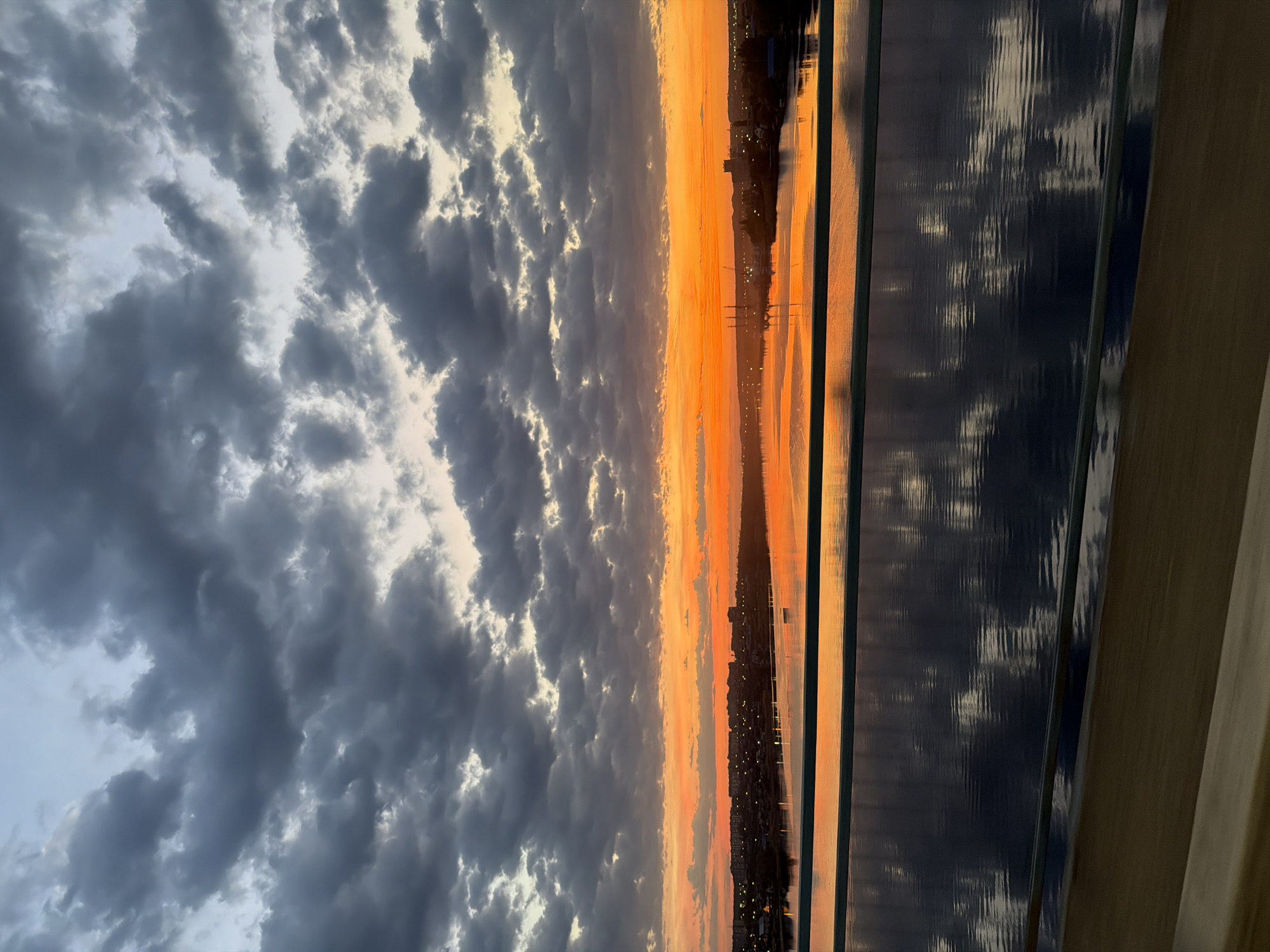 The image shows a sunset over Kamyshin, Russia. The sky is filled with dark, billowing clouds on the left, transitioning to a bright orange and yellow horizon. In the distance, the silhouette of the city skyline is visible, dotted with lights. The scene appears to be viewed through a window, as reflections are visible on the glass panels.